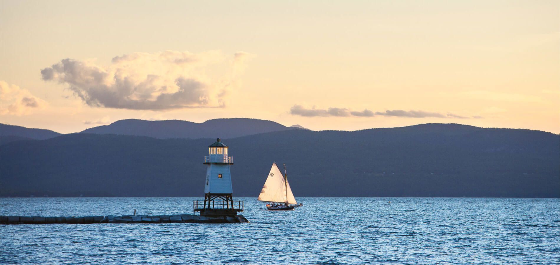 Sailboat passing a small white lighthouse on the lake at sunset, with dark mountains behind