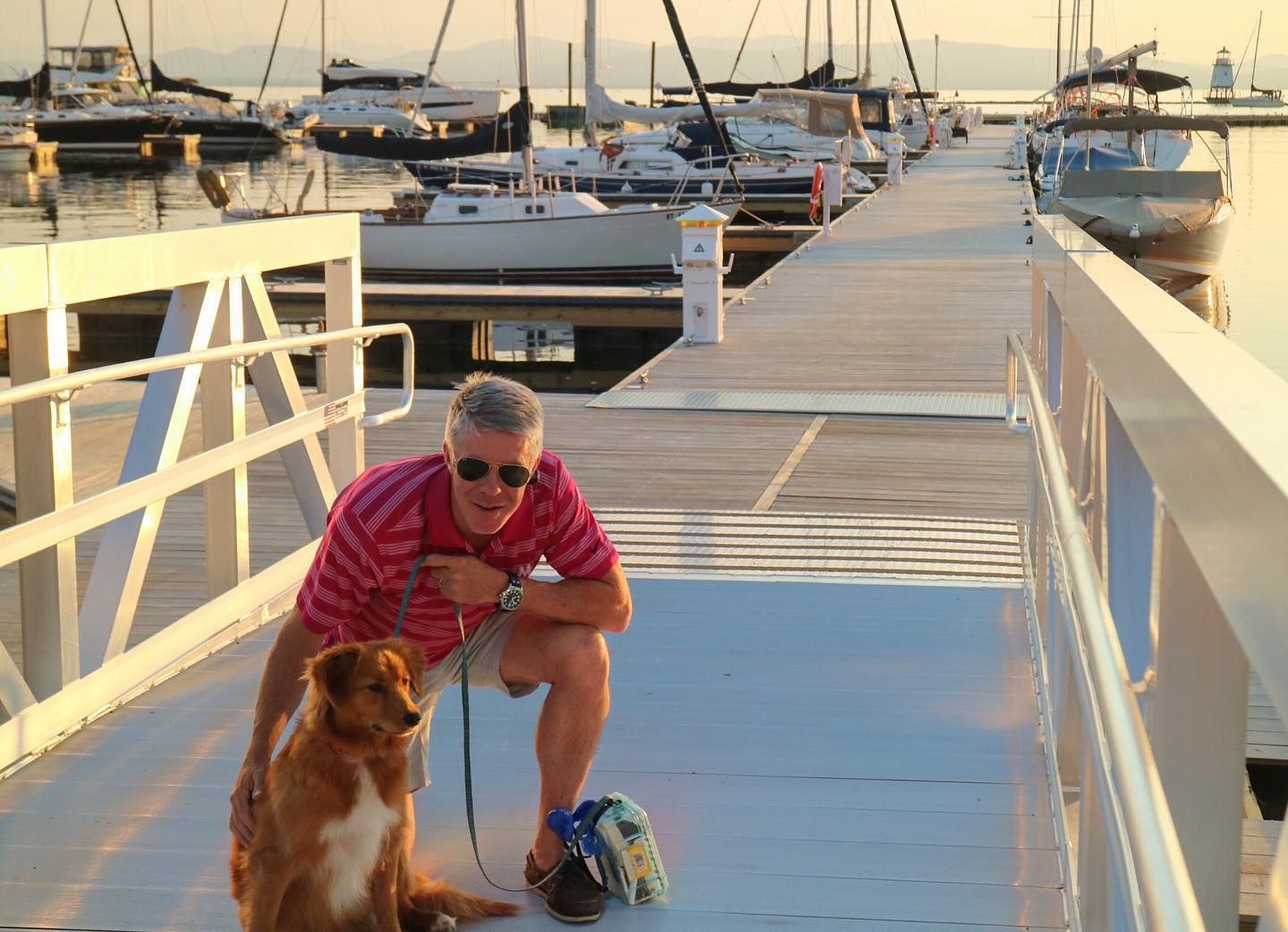 Person crouching with a dog on a floating dock at sunset, with sailboats moored behind