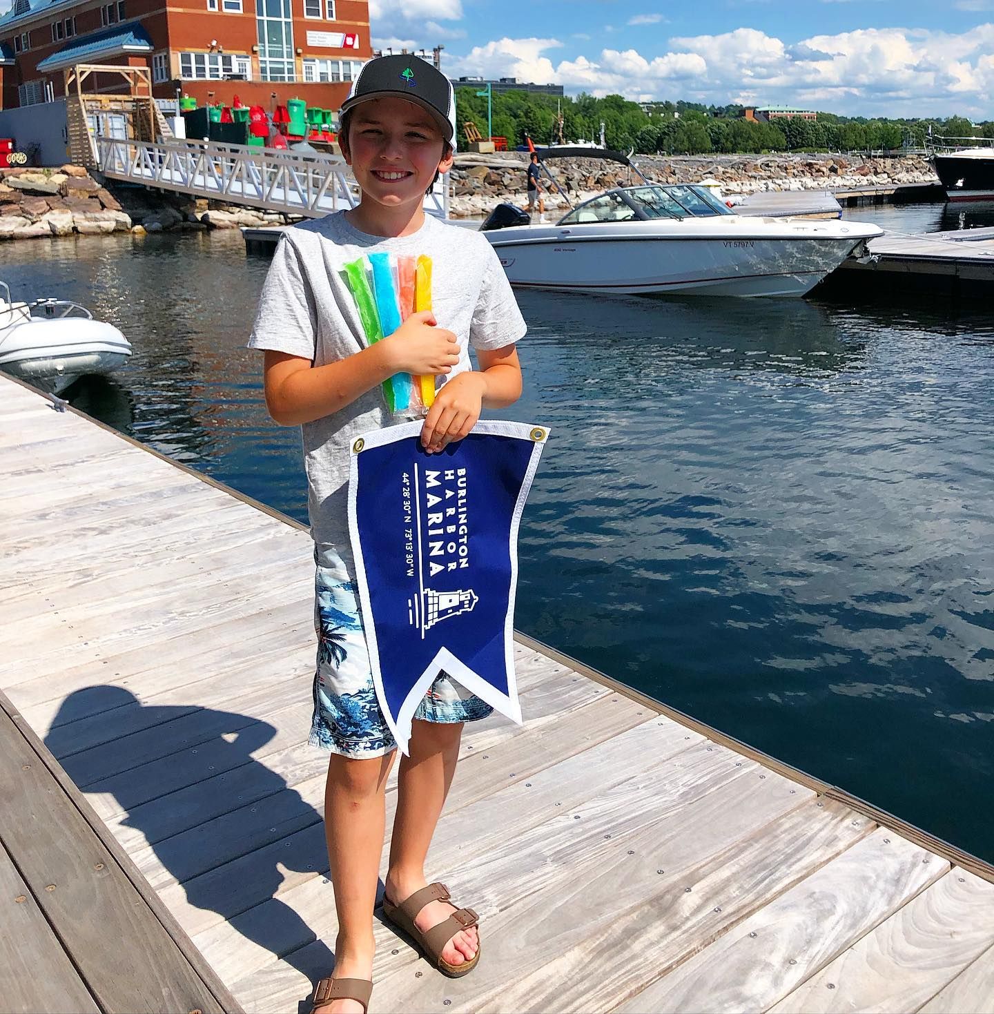 Child holding colorful ice pops and a Burlington Harbor Marina pennant on the dock, with boats behind