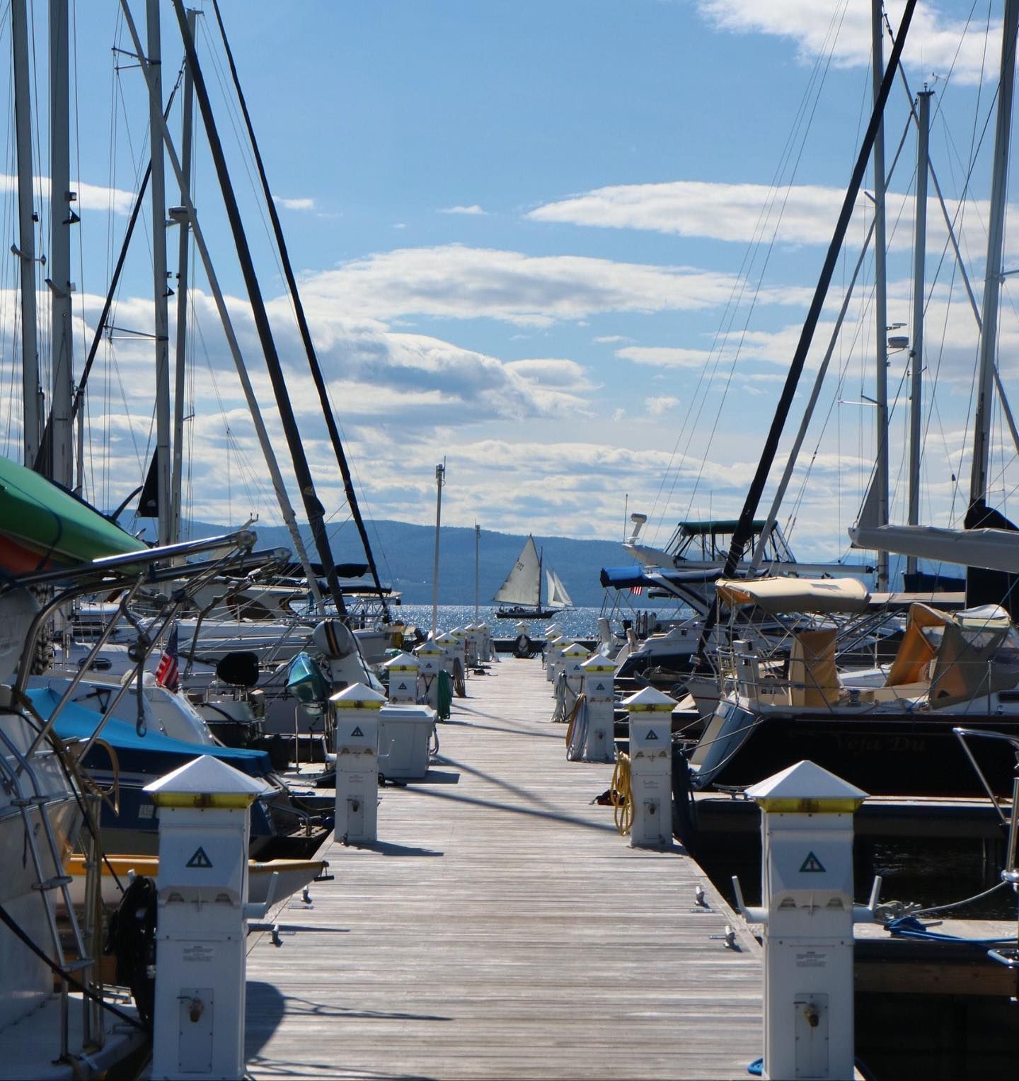 Sailboat masts and rigging framing a blue sky, with a small sailboat and mountains in the distance