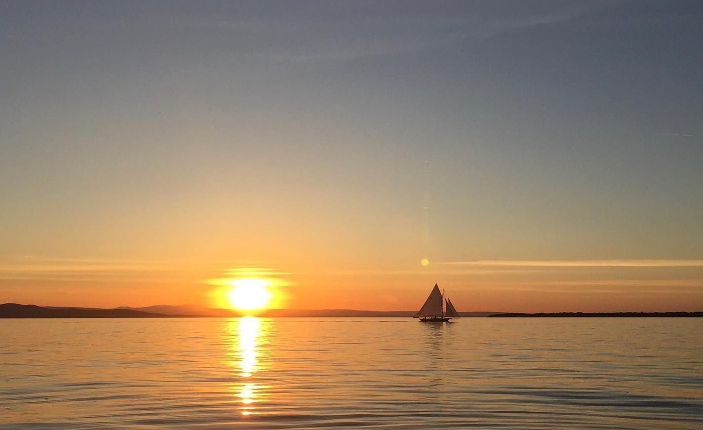 Sailboat on calm water at sunset, with the sun low on the horizon and its reflection on the lake