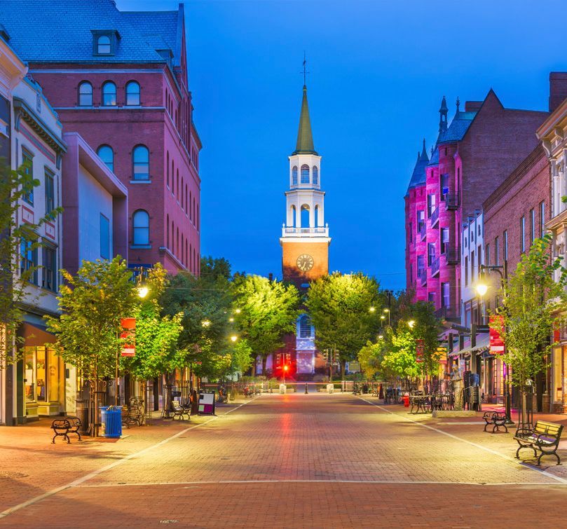 Historic brick buildings and a white church steeple with a clock, lit at dusk on a downtown street