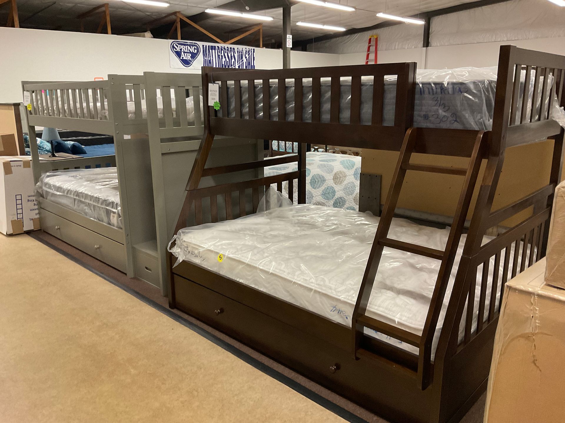 Two triple bunk beds in a warehouse. One is gray, the other brown, with ladders and mattresses.