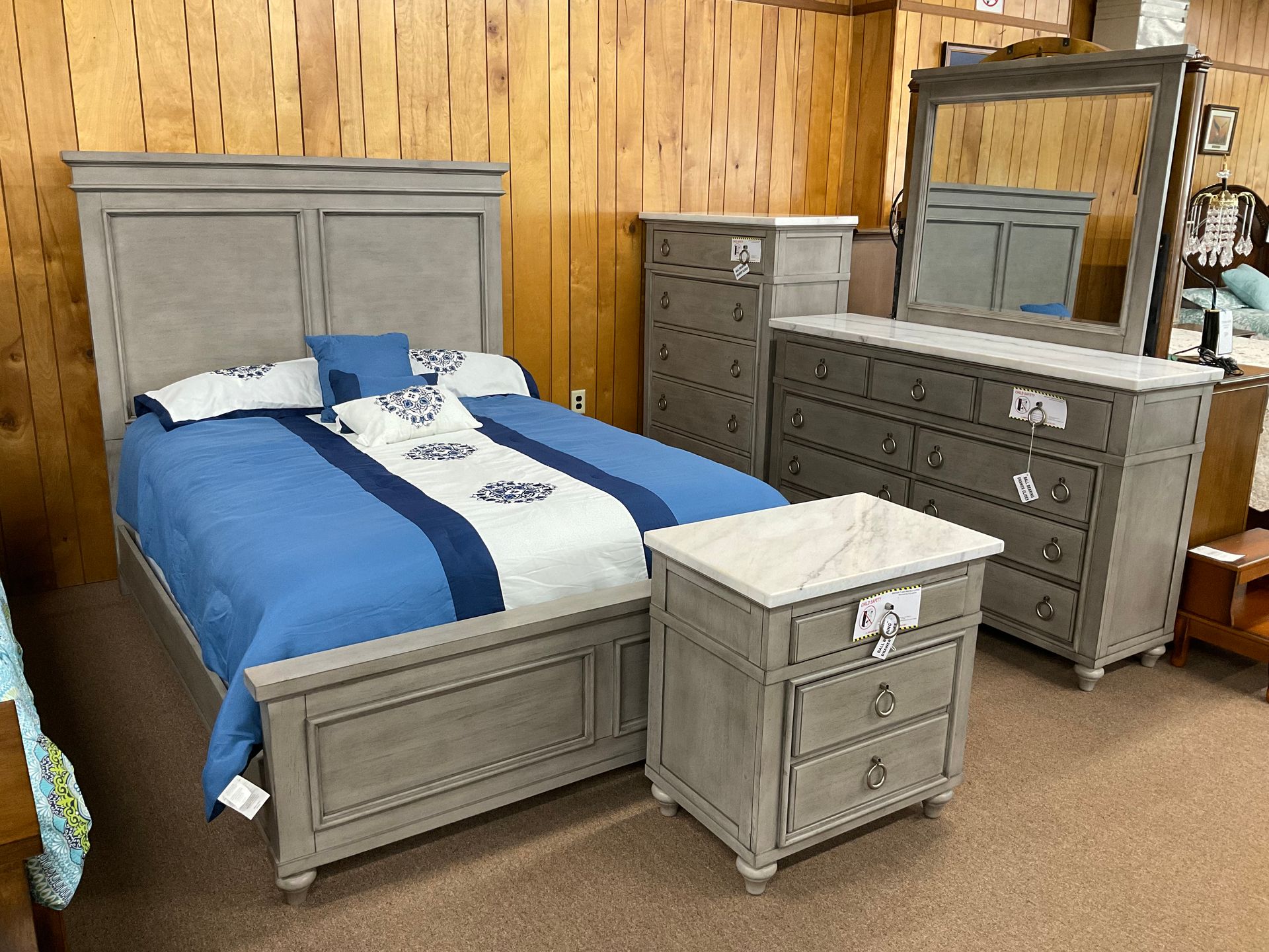 Bedroom furniture set: gray bed, dresser, chest, nightstand, and mirror against a wood-paneled wall.