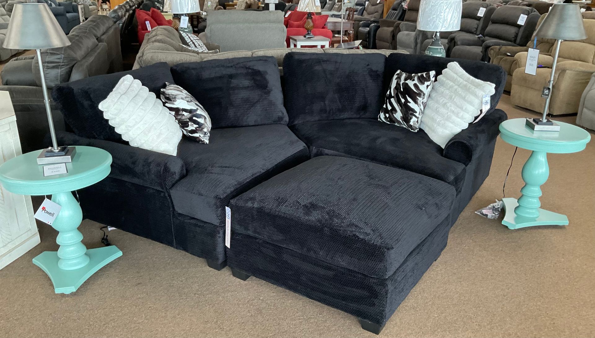Black sectional sofa with ottoman, teal side tables, lamps, and decorative pillows in a furniture store.