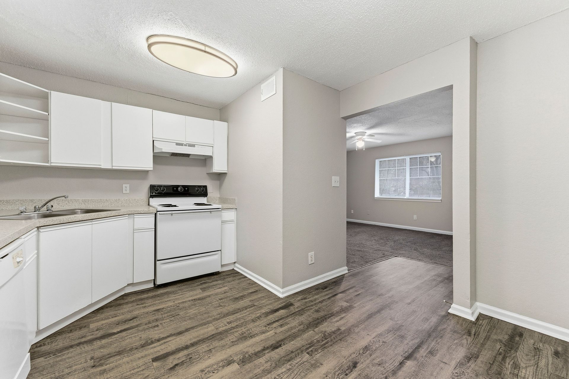 Empty kitchen with white cabinets, stove, and gray wood-look flooring opening into a carpeted living room.