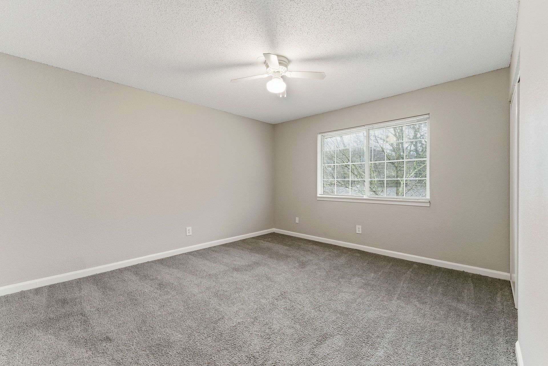 Empty beige bedroom with gray carpet, white ceiling fan, and window with blinds