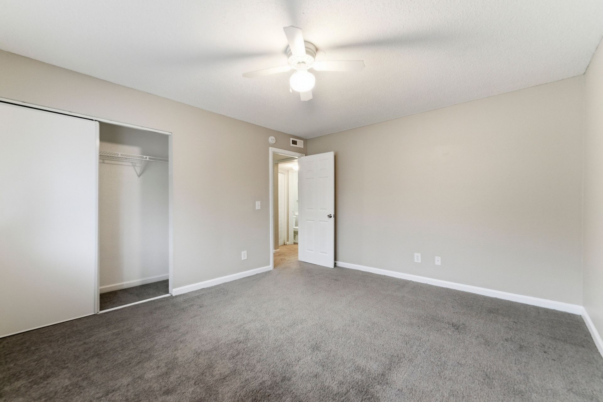 Empty beige bedroom with gray carpet, open closet, and ceiling fan pendant light
