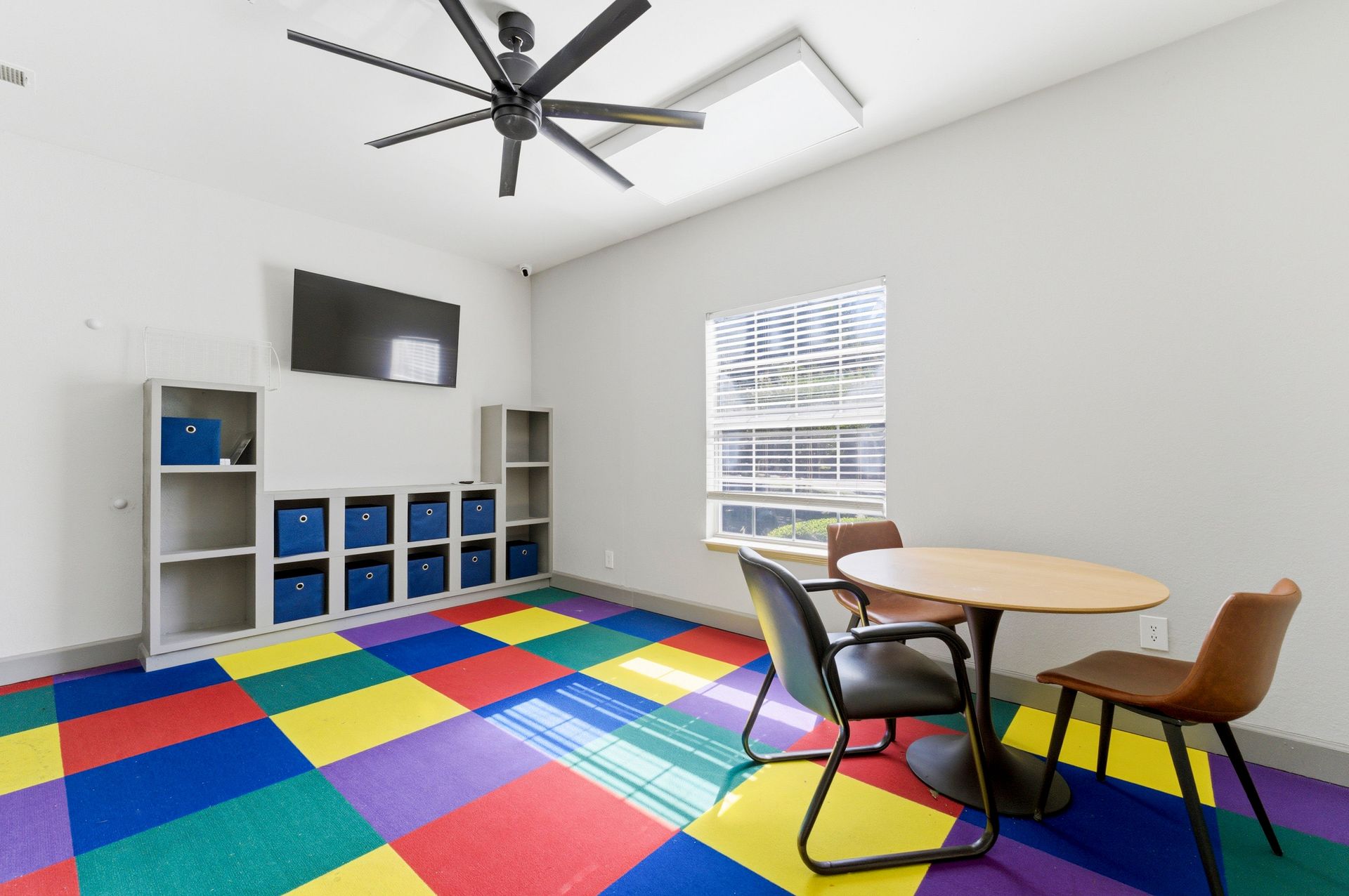 Colorful playroom with a rainbow foam floor, storage cubbies, round table, chairs, and wall-mounted TV.