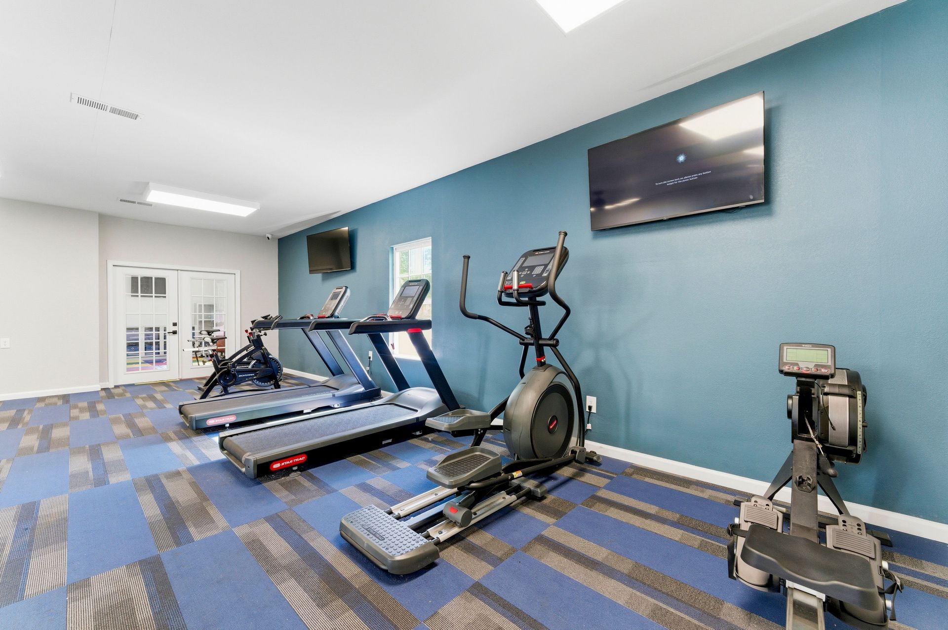 Gym room with treadmills, elliptical, rowing machine, and TV on a blue wall