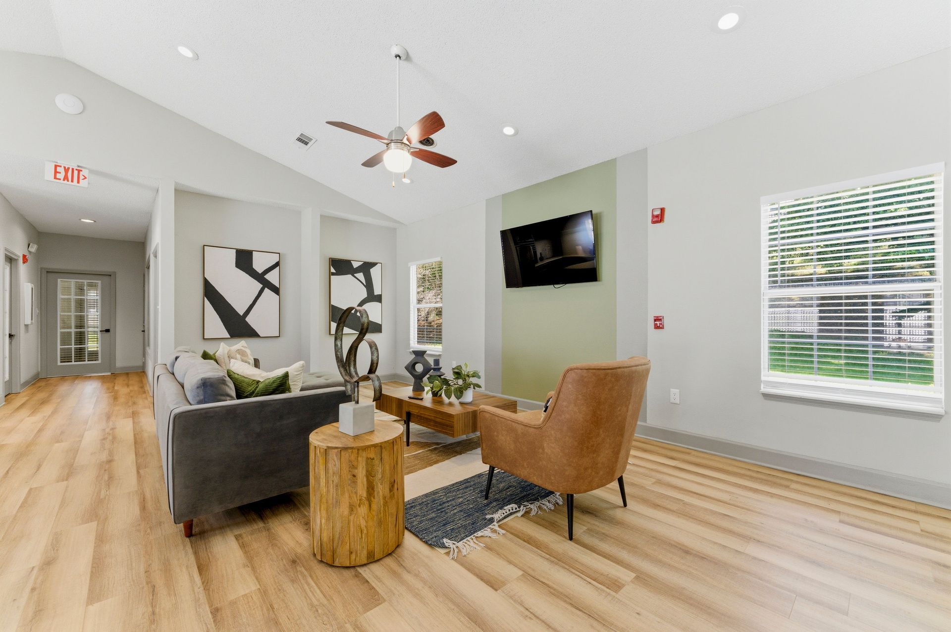 Bright modern living room with gray sofa, tan chair, wall-mounted TV, wood floors, and large windows.