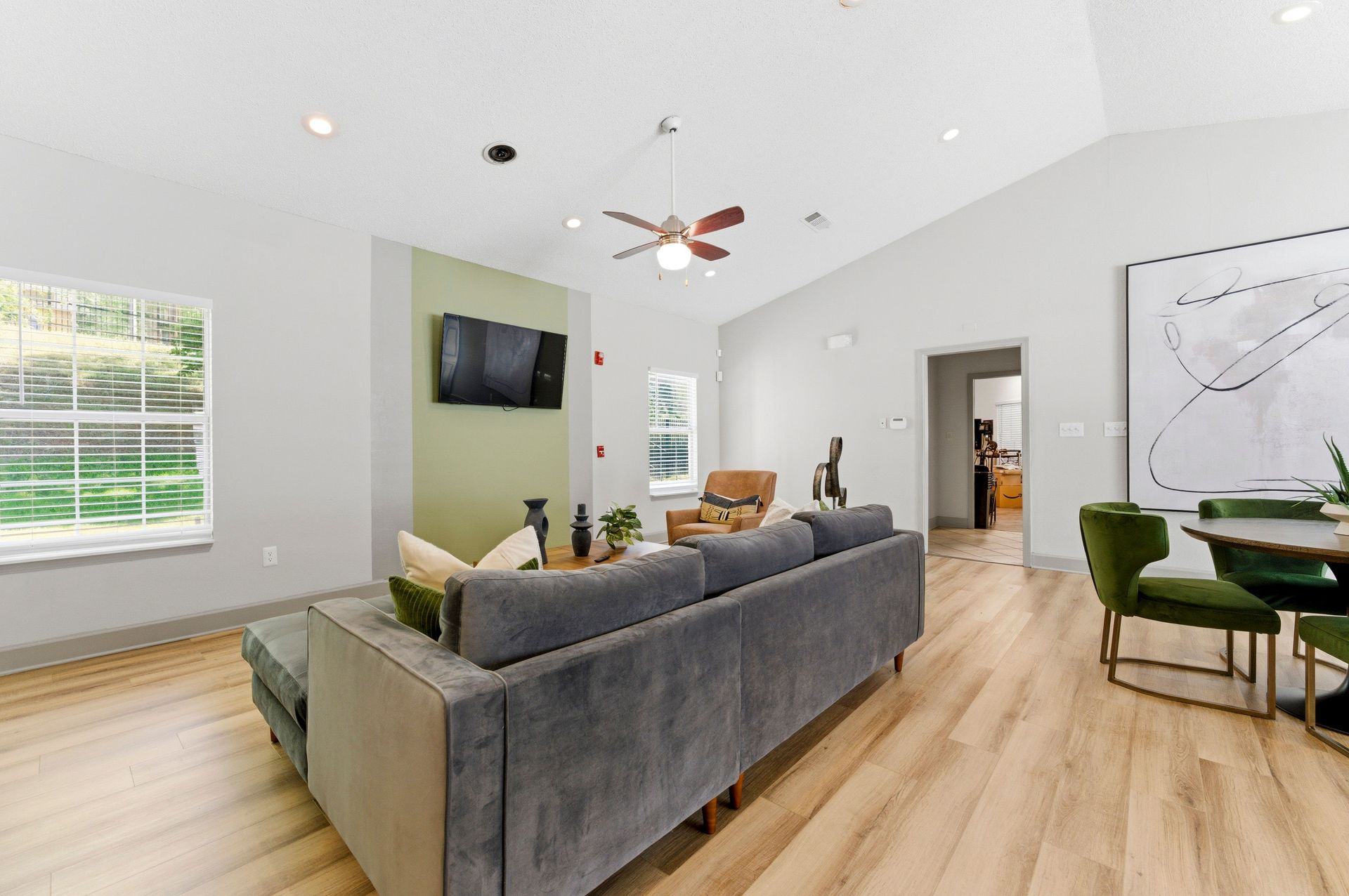 Modern open-plan living room with gray sofa, wall TV, wood floors, and dining area by a window.