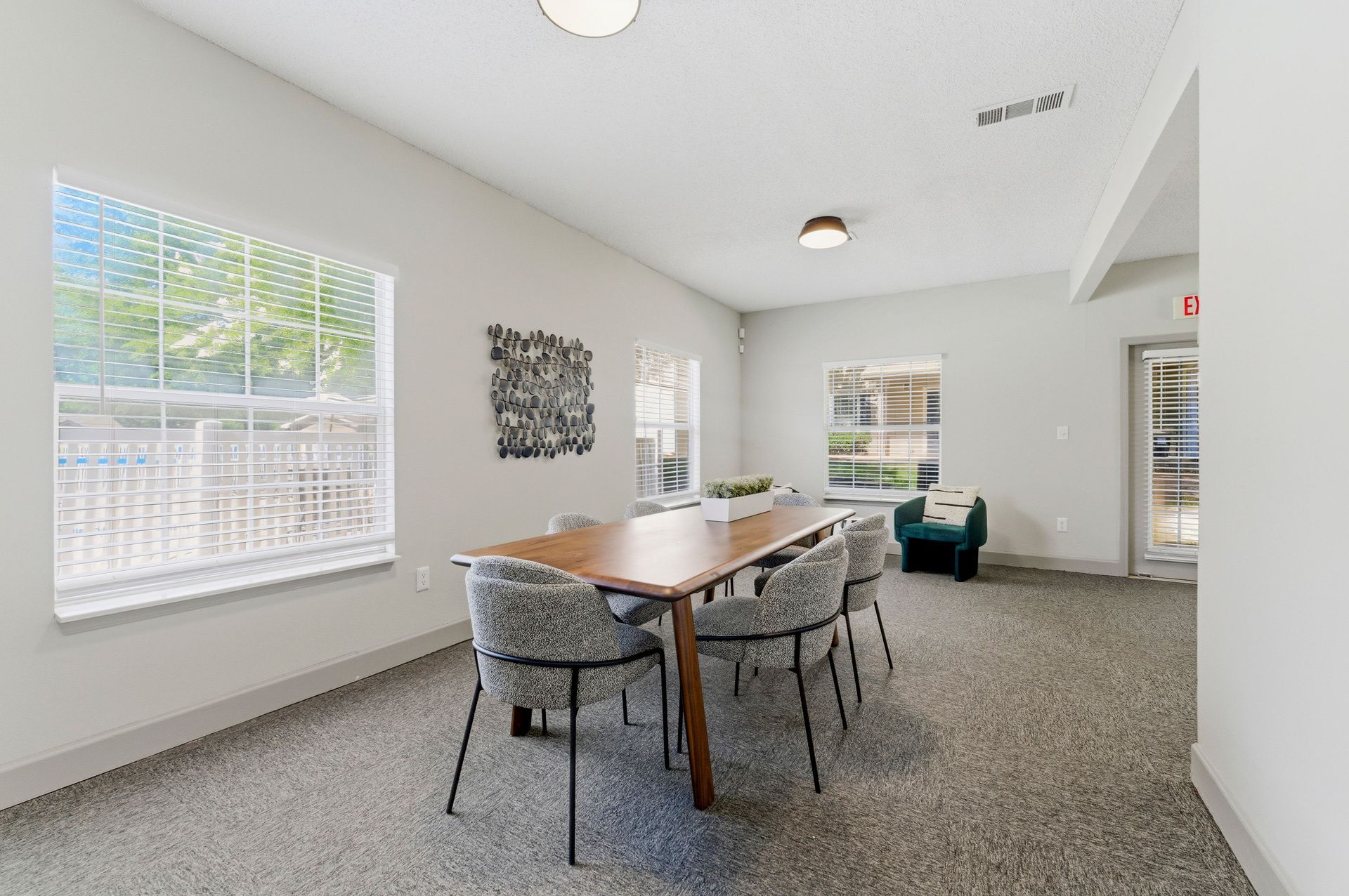 Bright meeting room with a long wooden table, patterned chairs, windows, and a teal armchair.