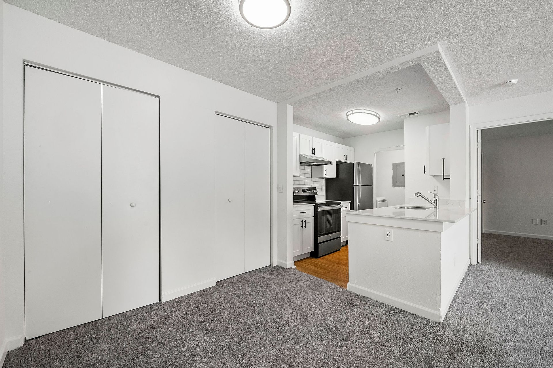 Empty apartment living area with carpet, white walls, closet doors, and a small kitchen nook.