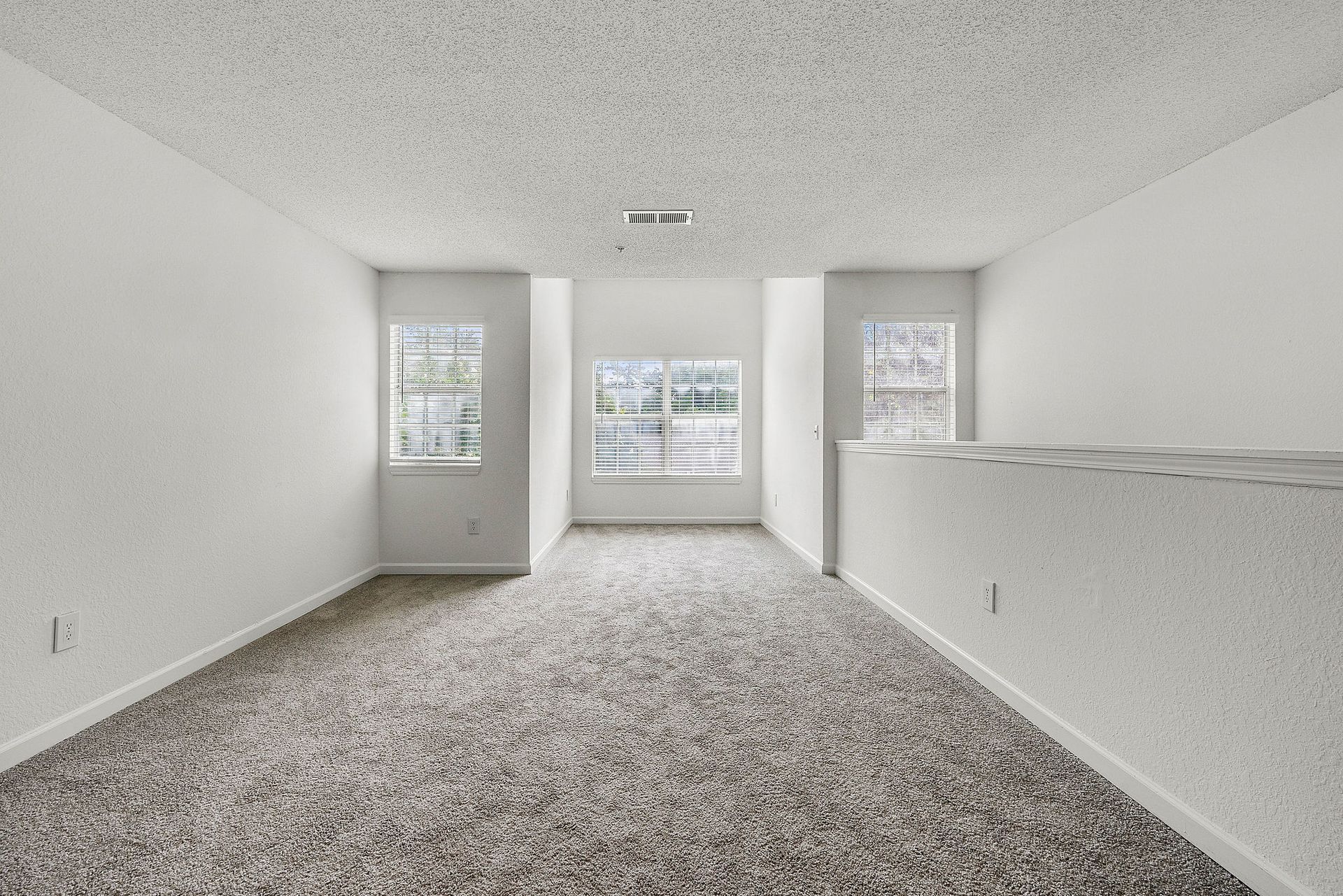 Empty carpeted living room with white walls and windows overlooking a railing