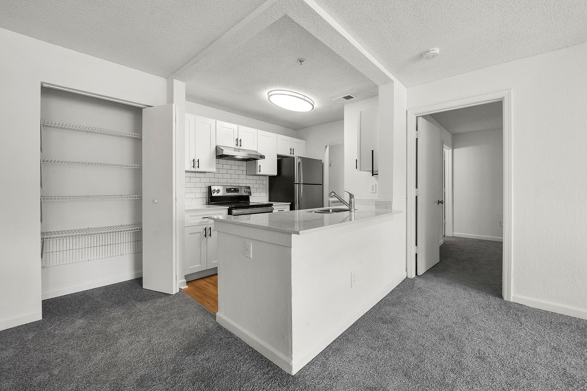 Empty apartment kitchen and hallway with gray carpet and white walls, viewed from the living area.