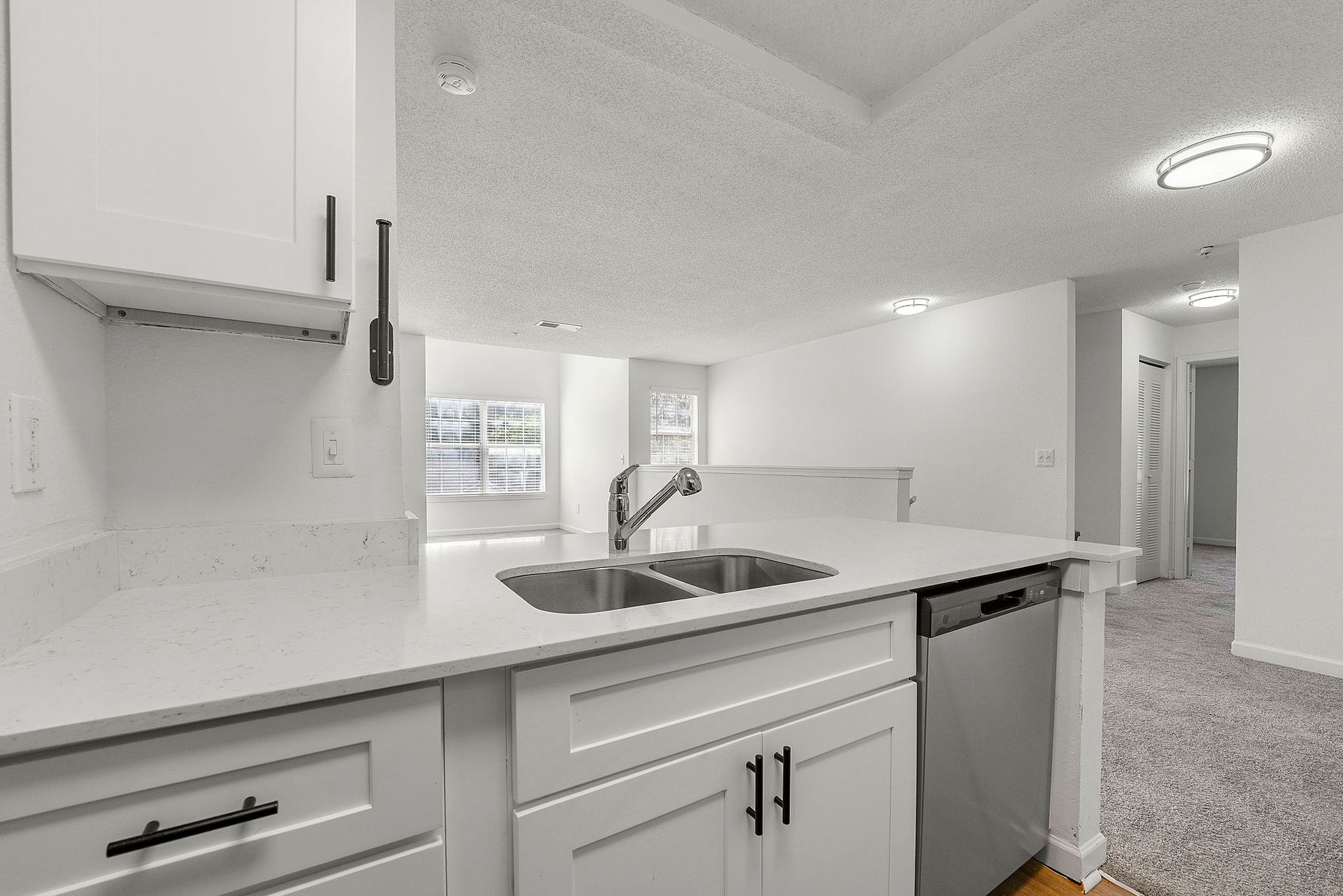 Bright white kitchen with sink, cabinets, and a view into an adjoining room with carpet.