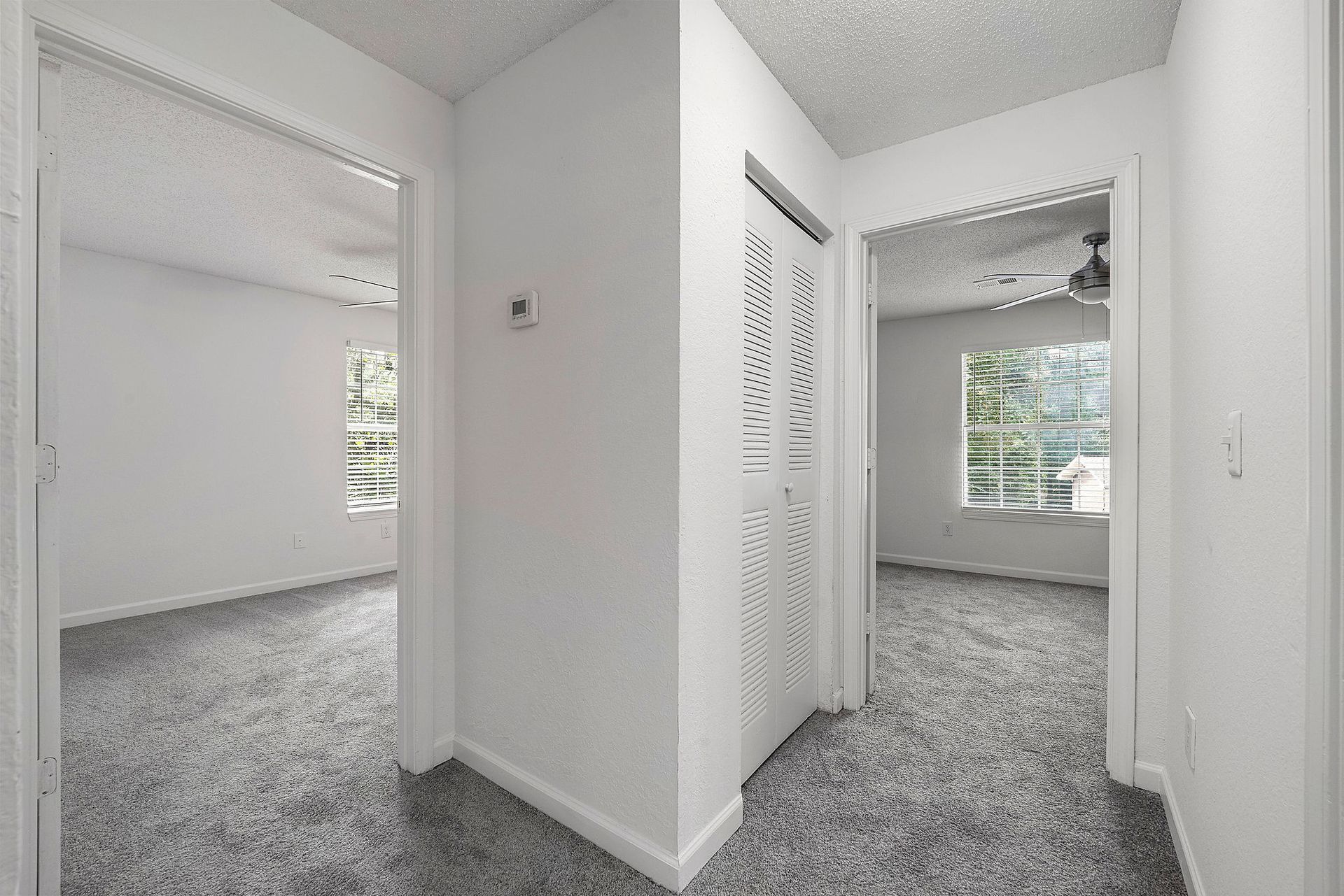 Empty white hallway with carpet, opening into two bright rooms with windows and closet doors