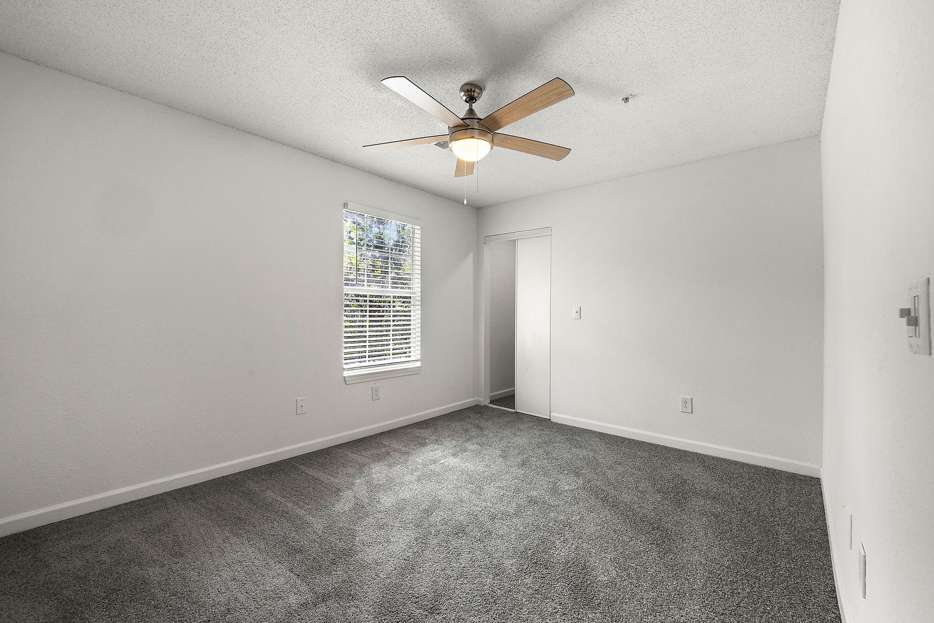 Empty carpeted bedroom with white walls, ceiling fan, and a window with blinds