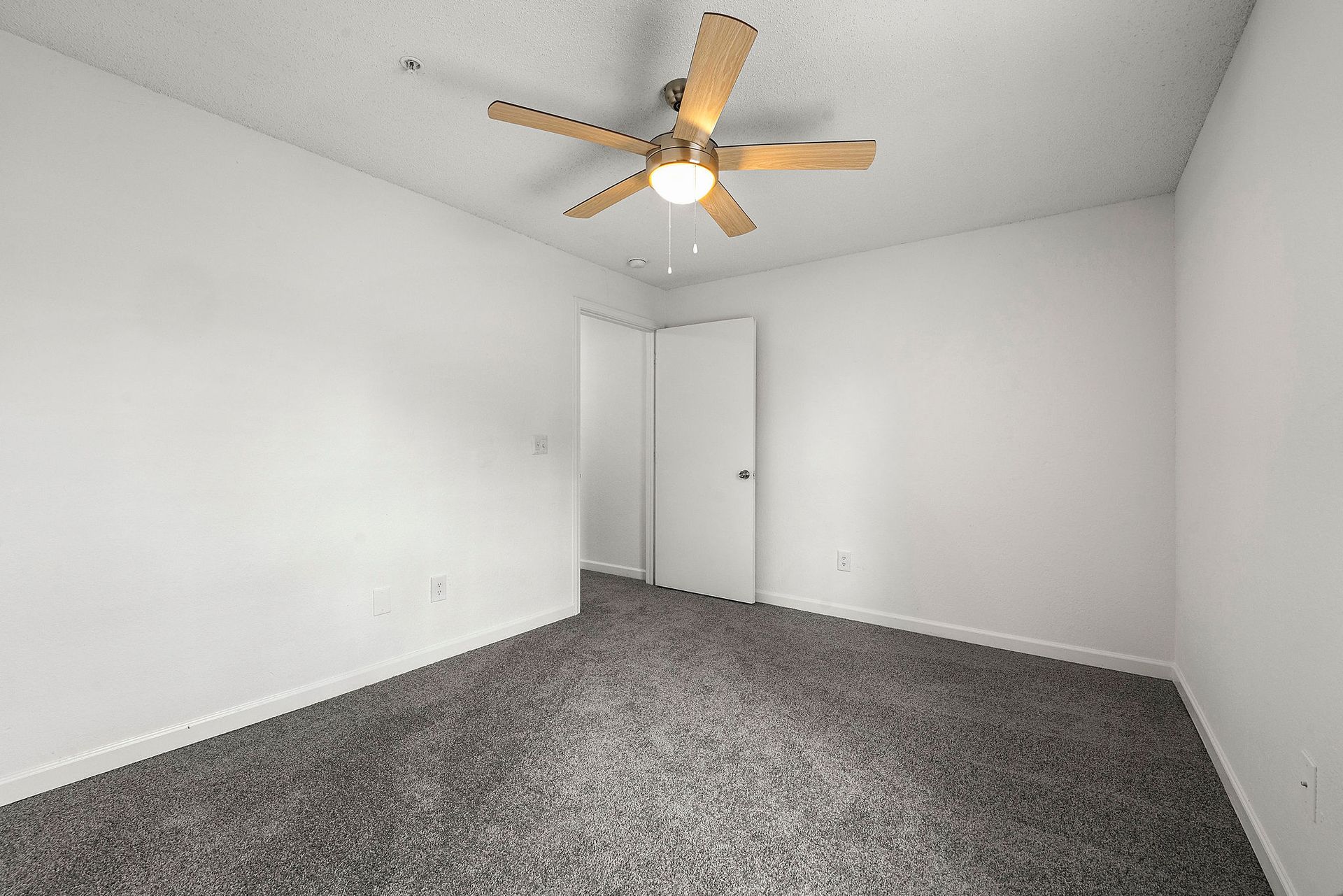 Empty white bedroom with gray carpet and a ceiling fan, viewed toward a closed closet door.