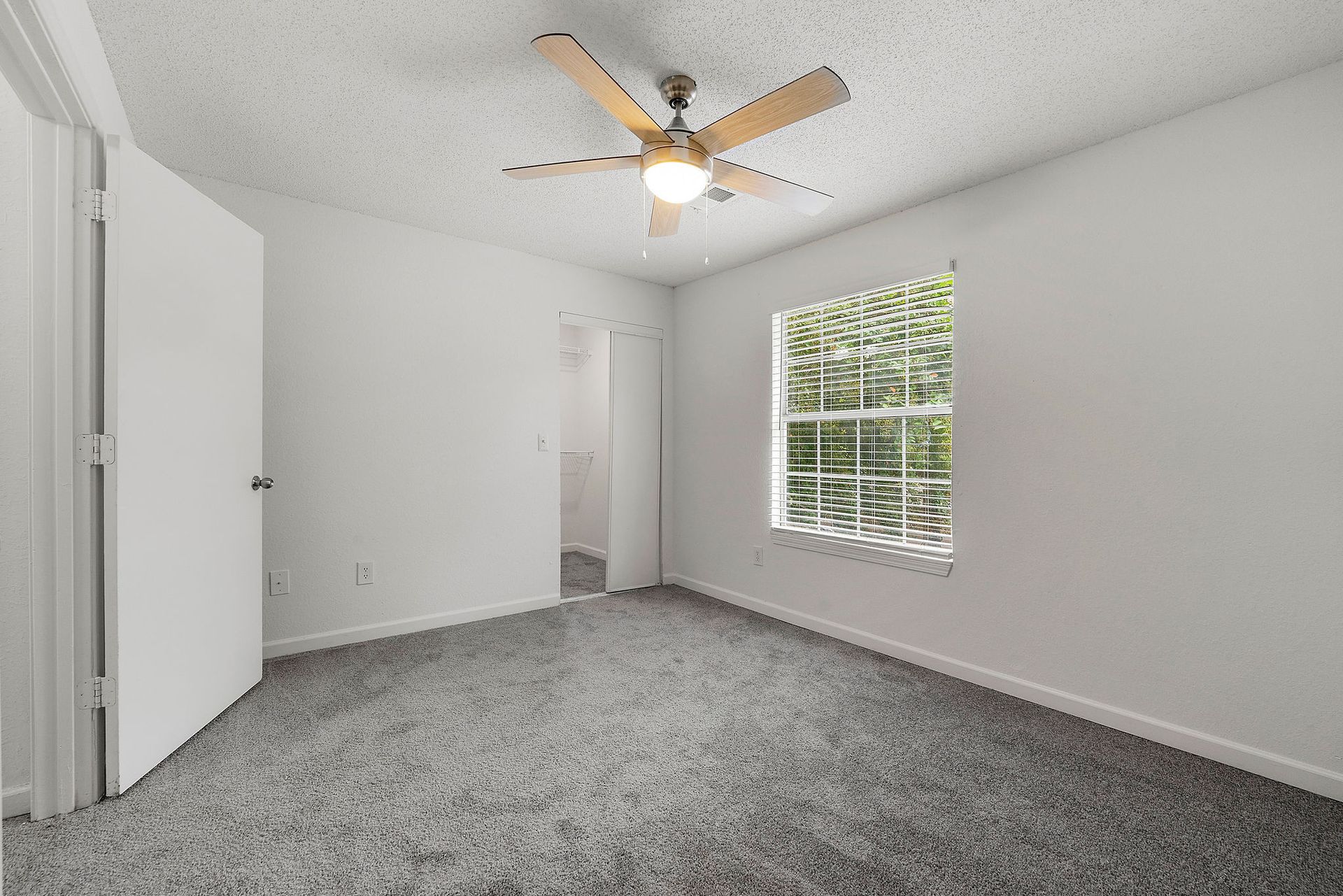 Empty carpeted bedroom with white walls, ceiling fan, closet doors, and a window with blinds.