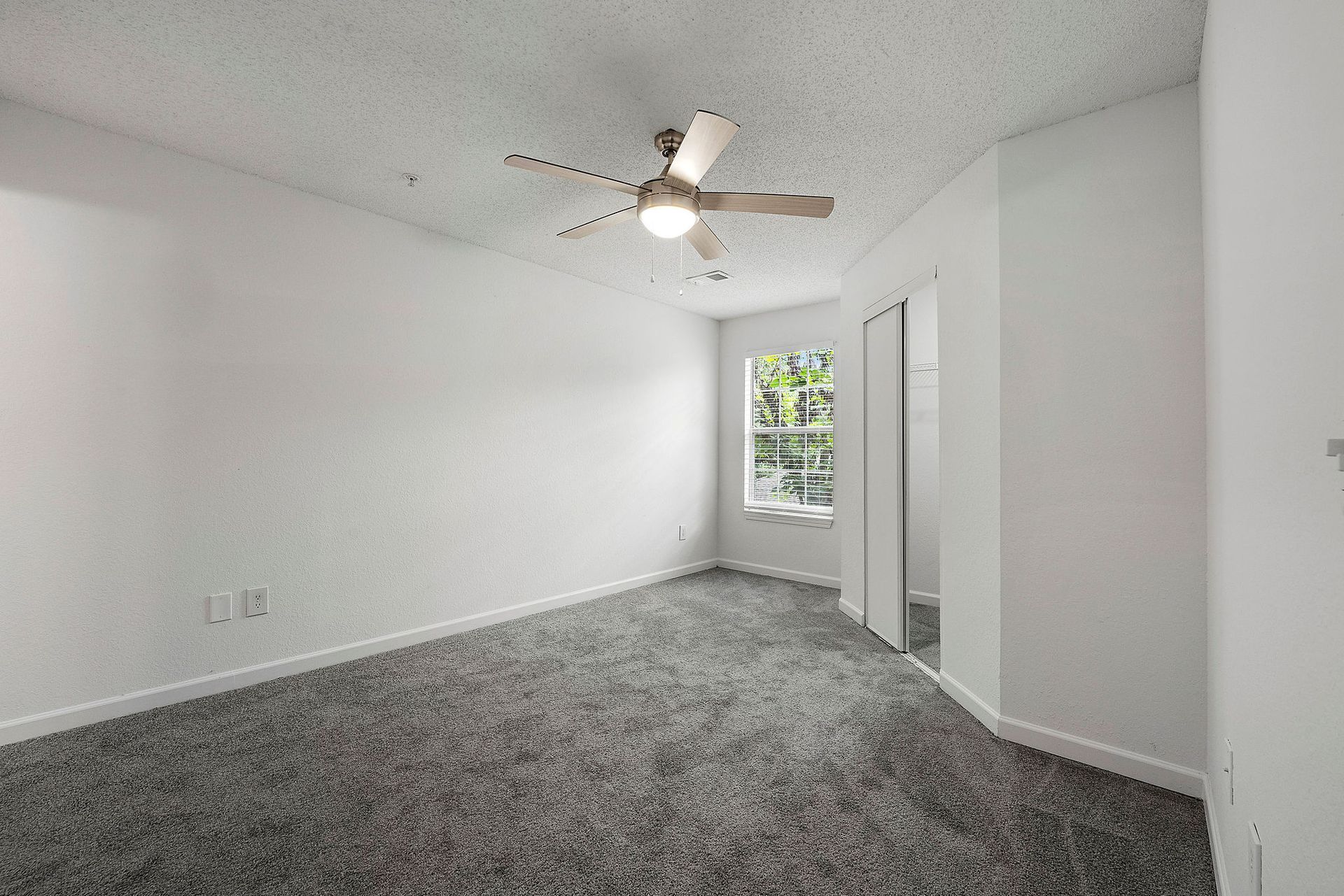 Empty white bedroom with gray carpet, ceiling fan, and window light.