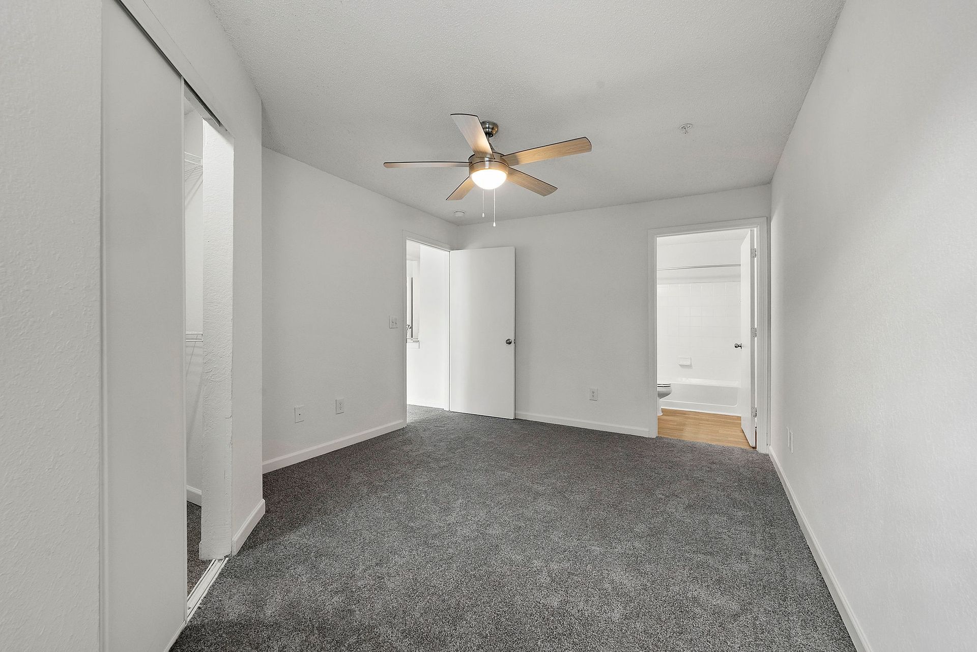 Empty carpeted bedroom with white walls, ceiling fan, and open closet and doorway