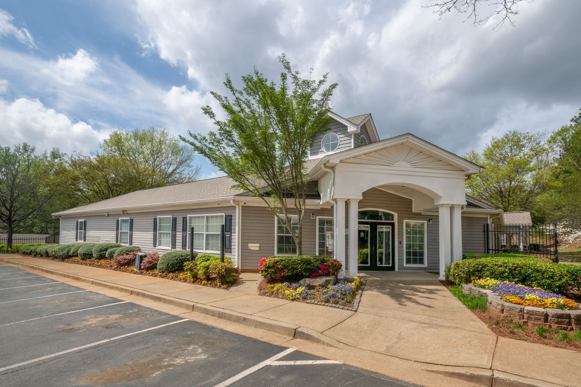 A small building with a covered entrance, flower beds, and a parking lot under a cloudy sky