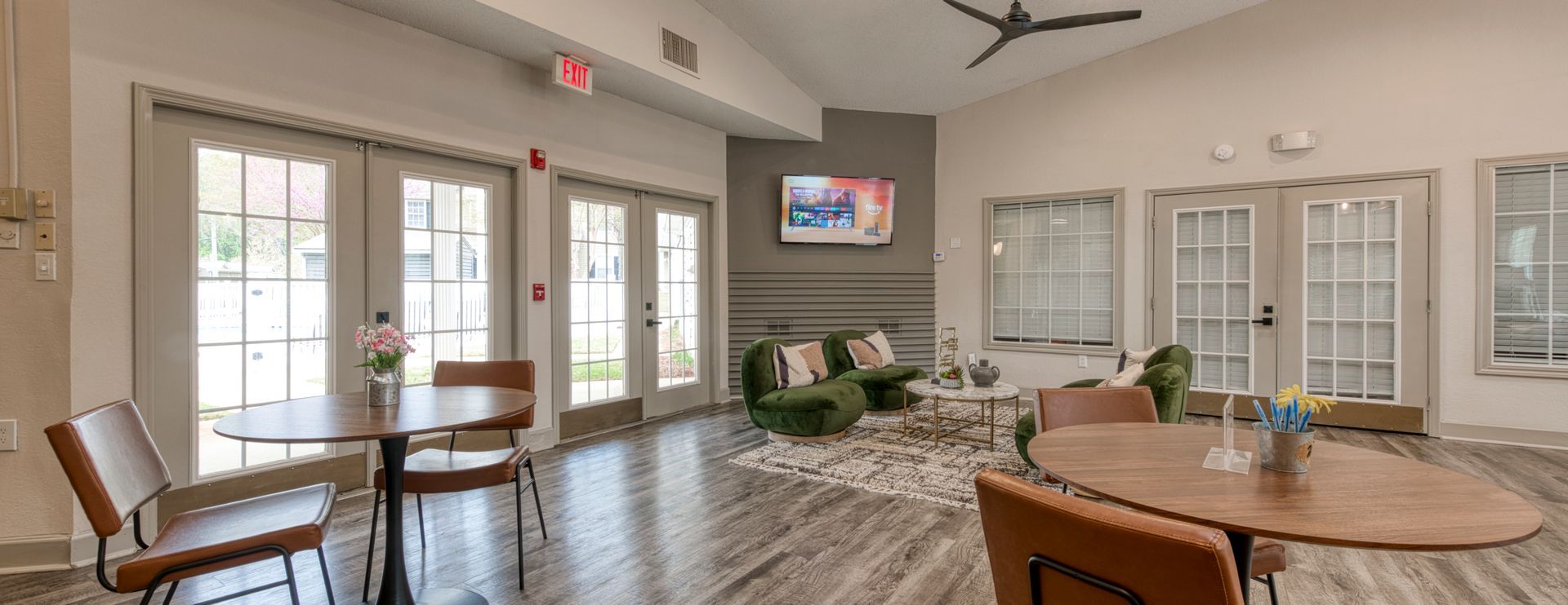 Bright lobby with tables, chairs, glass doors, potted plants, and a wall-mounted TV.