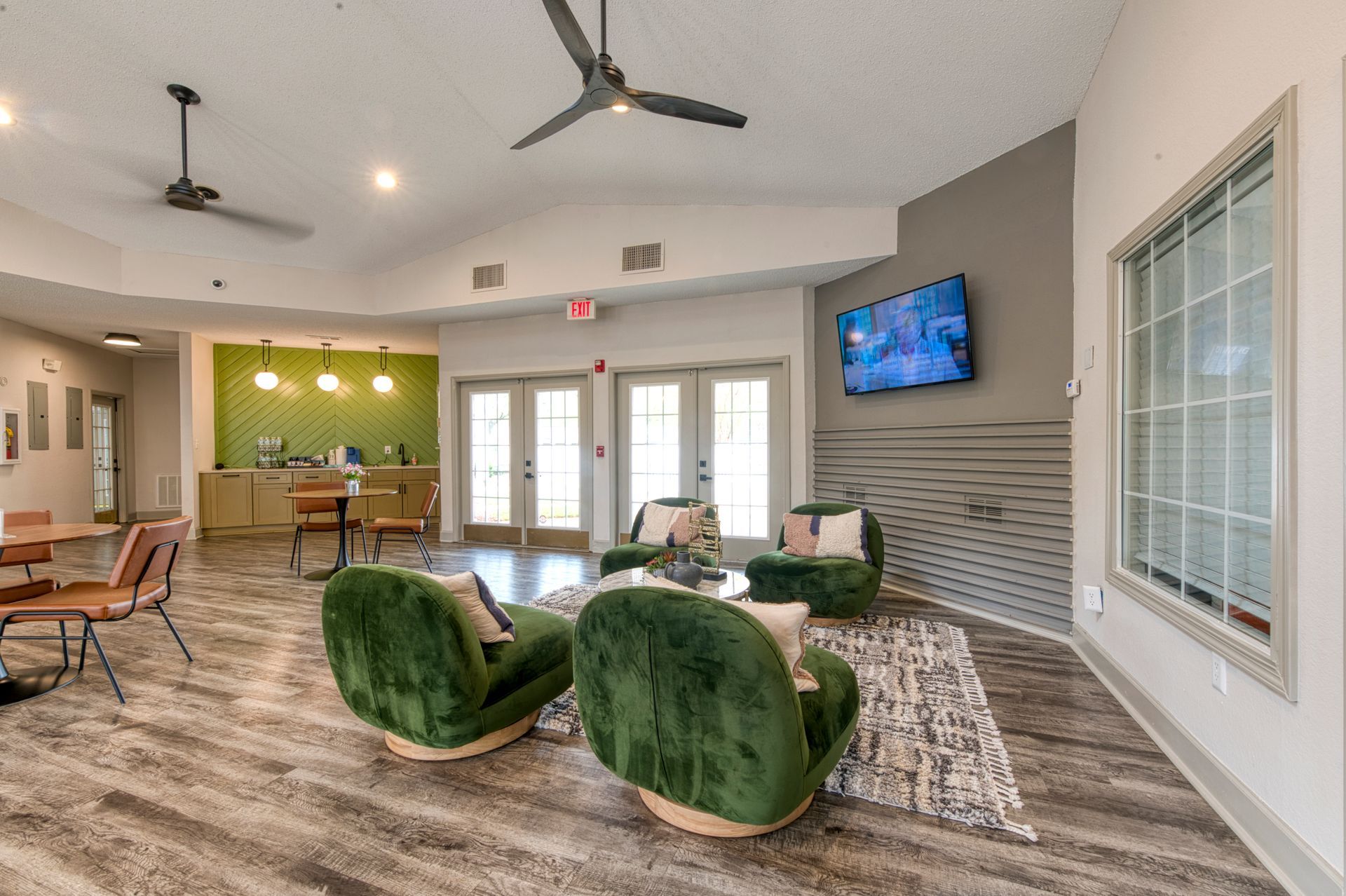 Modern apartment lounge with green chairs, wall-mounted TV, ceiling fans, and a kitchen bar in the background