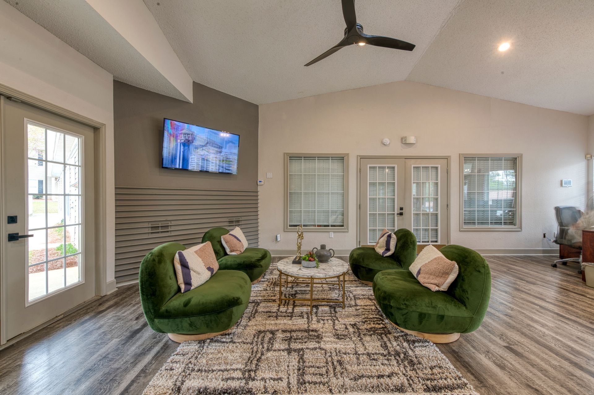 Modern living room with green chairs, patterned rug, wall TV, and large windows under a ceiling fan