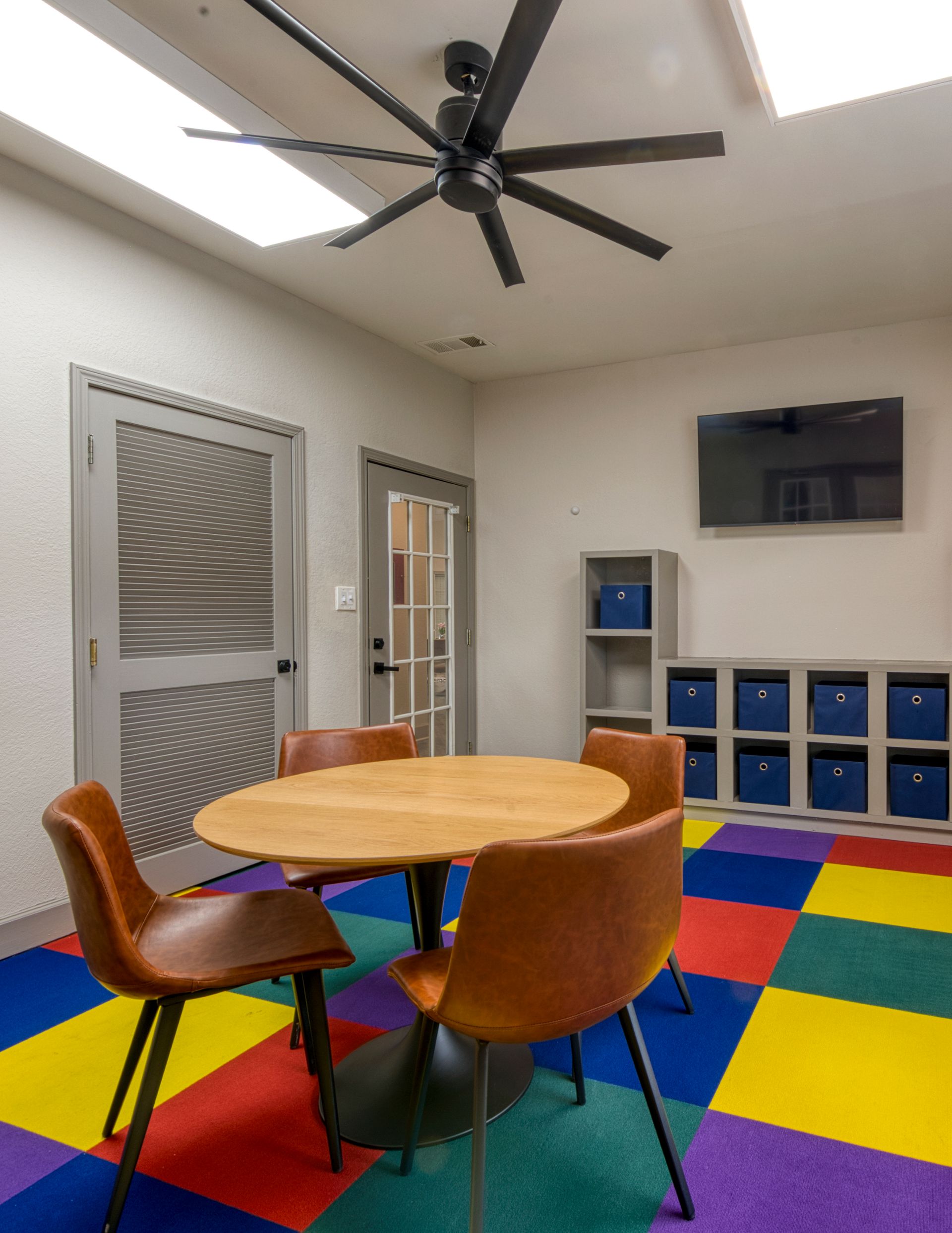 Colorful playroom with round table, chairs, wall-mounted TV, cubbies, and multicolor foam floor tiles.