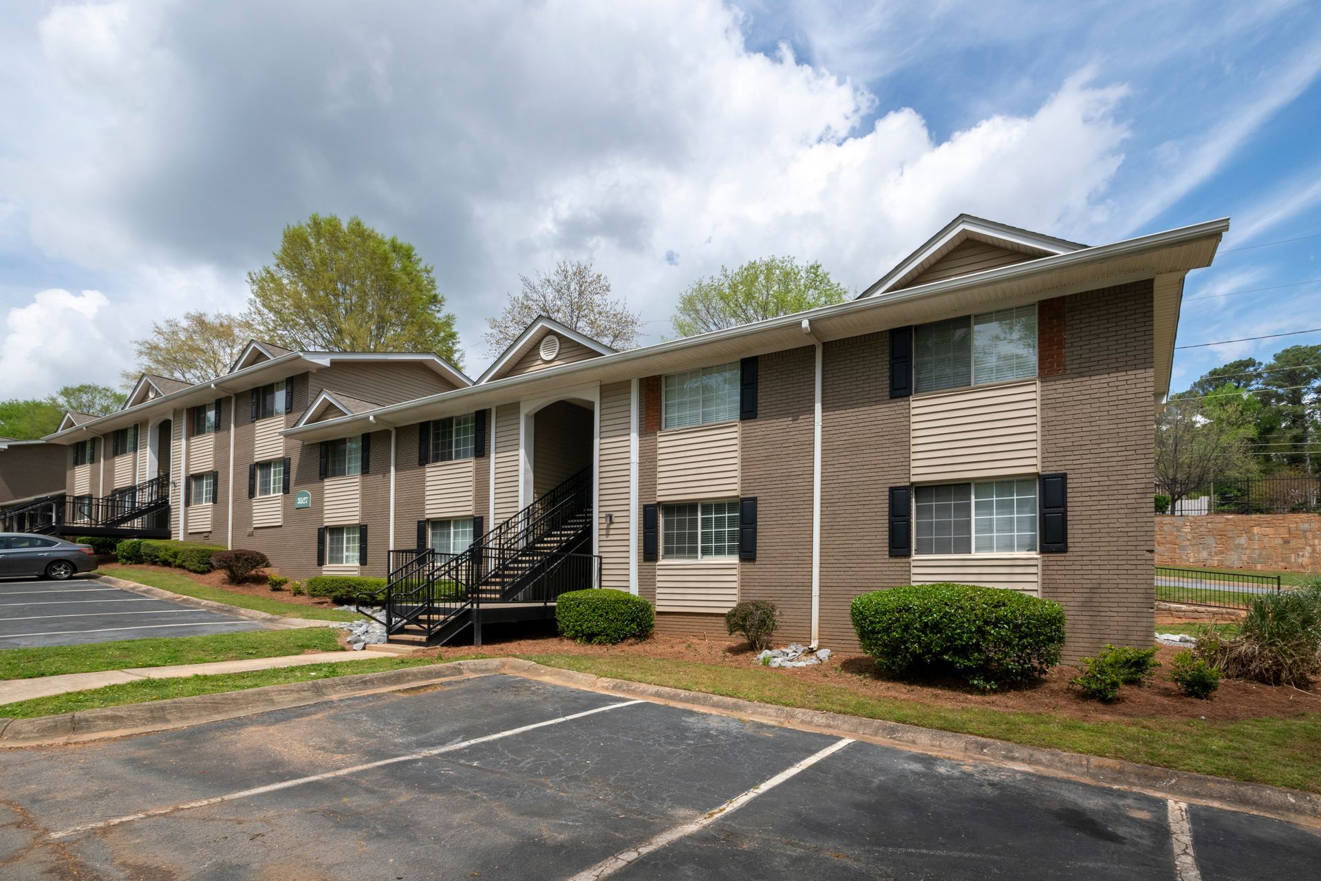 Row of tan brick apartment buildings with parking lot and trees under a cloudy sky