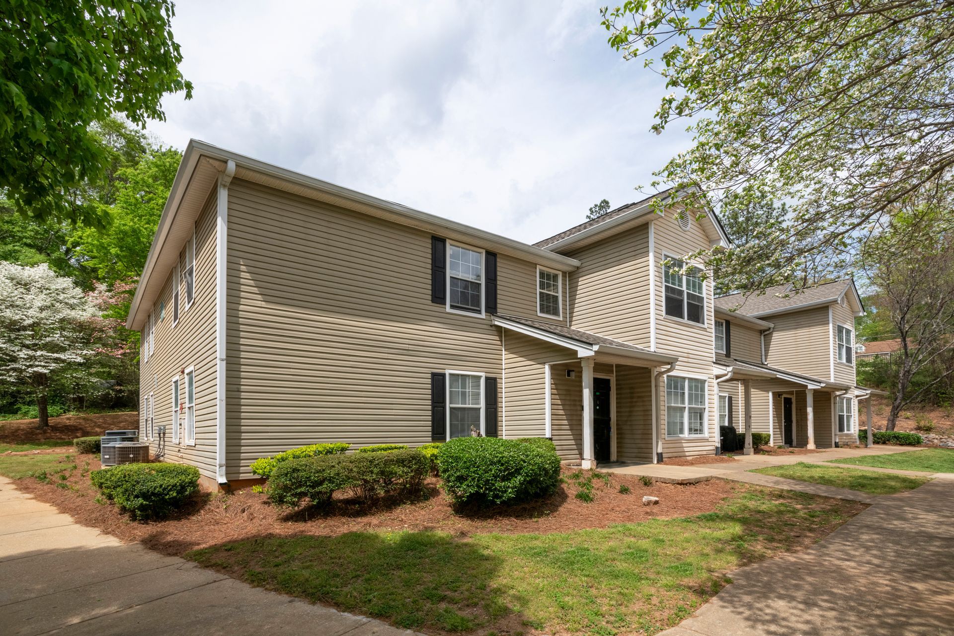 Tan apartment building with white trim and shrubs along a tree-lined walkway