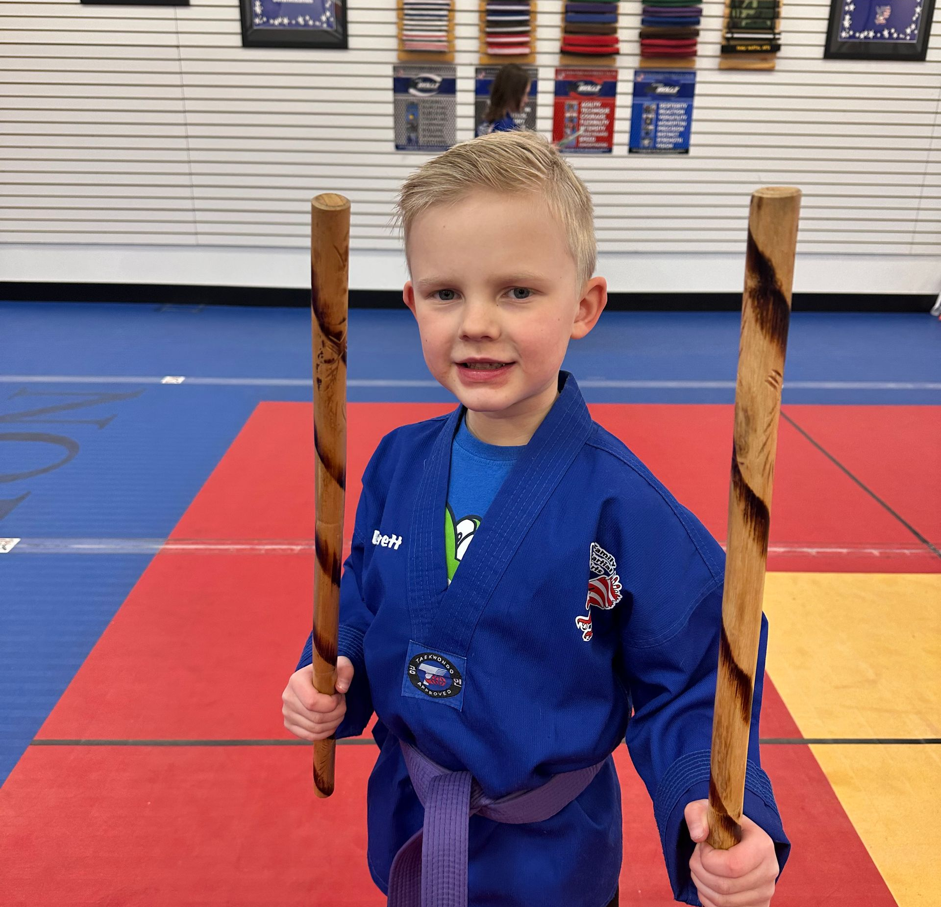 Smiling martial artist in white protective gear and helmet, standing on a mat in a gym.
