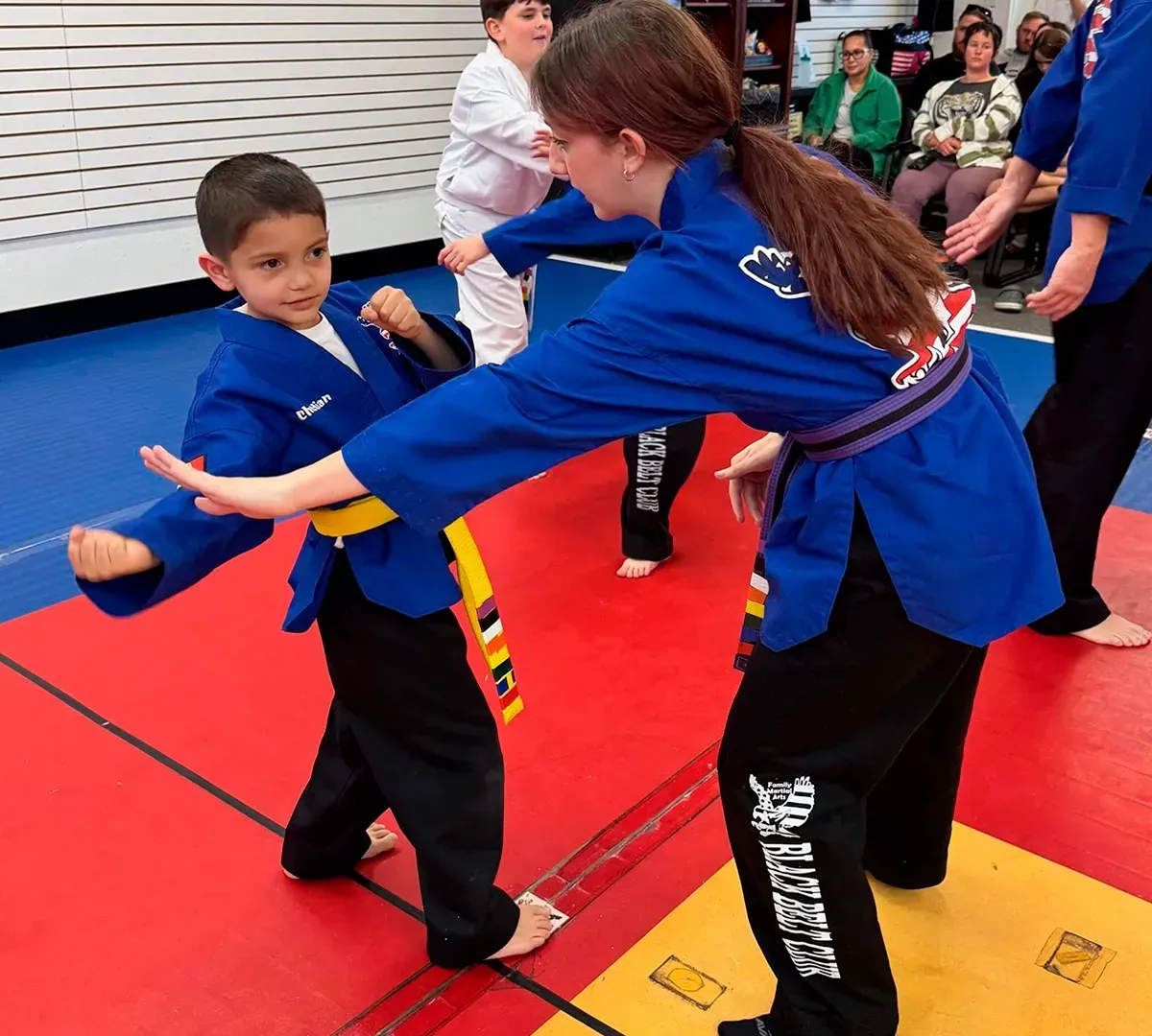 Martial arts class with two students practicing a move on red mats, instructor assisting nearby