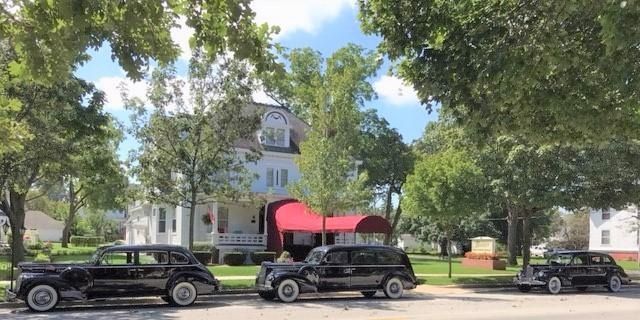 Three black vintage cars parked in front of a white house with a red awning and trees.