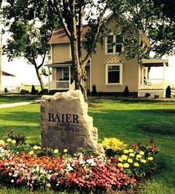 Stone sign reading “BAIER” in a flower bed in front of a yellow house with trees and lawn