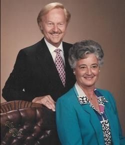 Studio portrait of two smiling adults, one seated in a teal jacket and one standing behind in a black suit