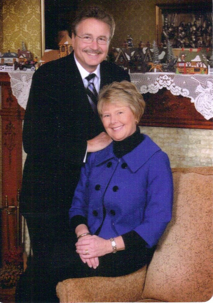 A smiling couple poses indoors by a decorated fireplace, with the woman seated in a blue coat.