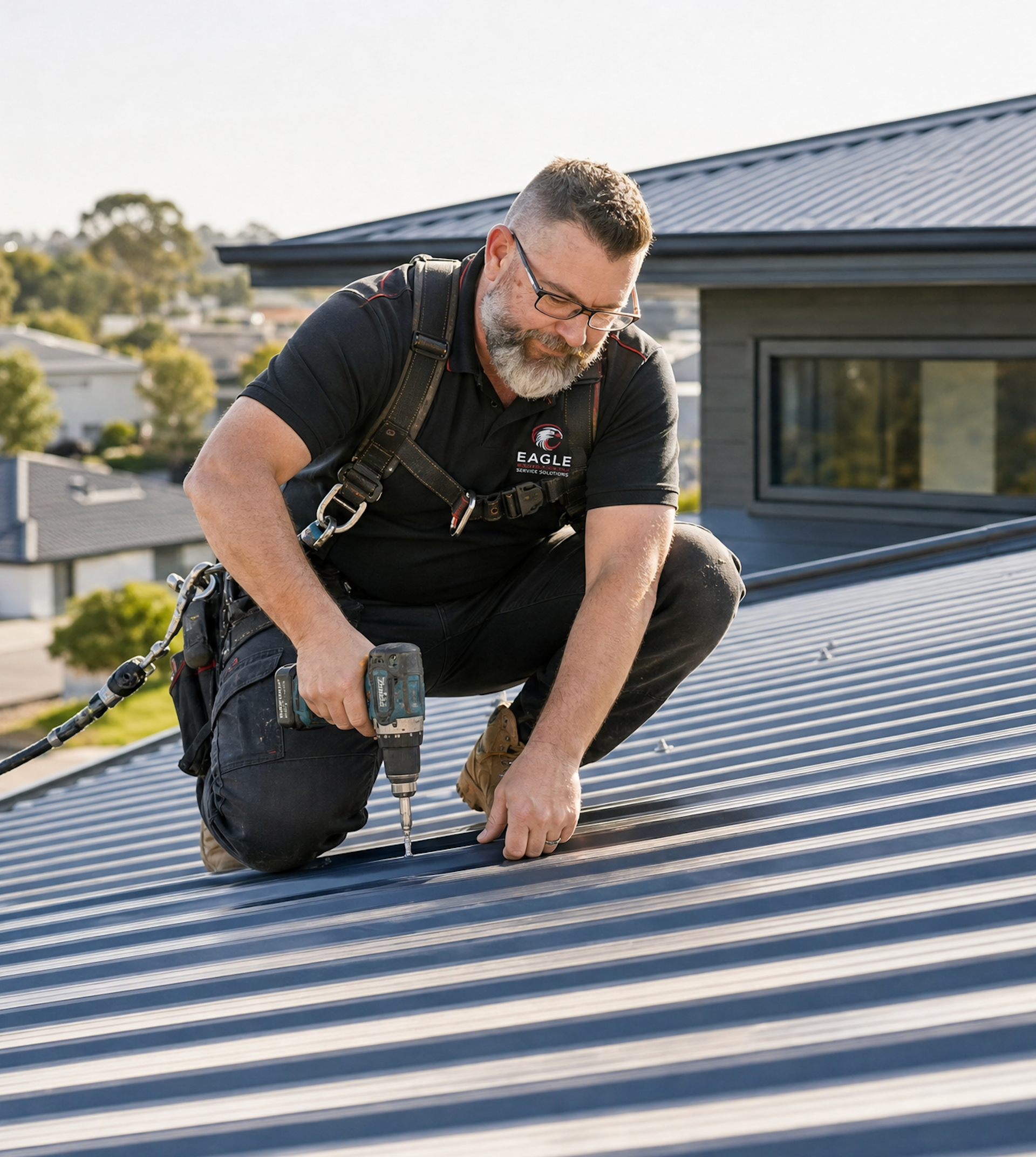 Worker kneels on a metal roof, using a power drill near a house window.