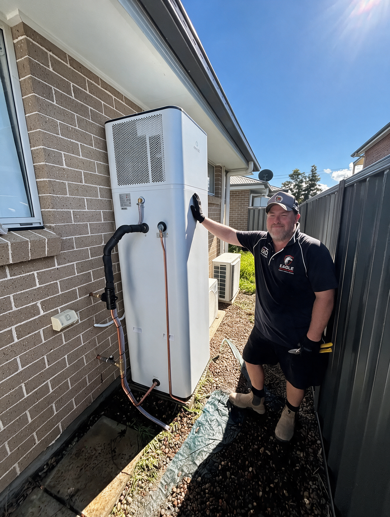 Man standing beside a large white outdoor air-conditioning unit next to a brick house.