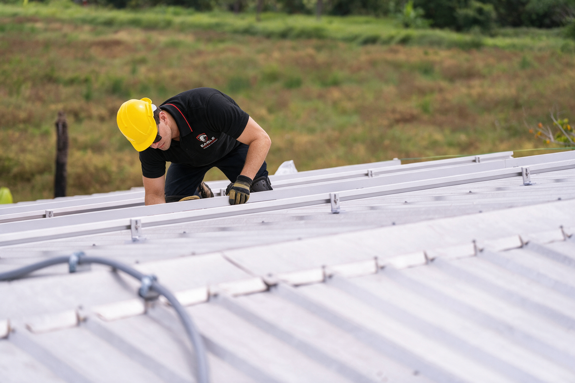 Worker in a yellow hard hat crouches on a metal roof, inspecting or repairing it.