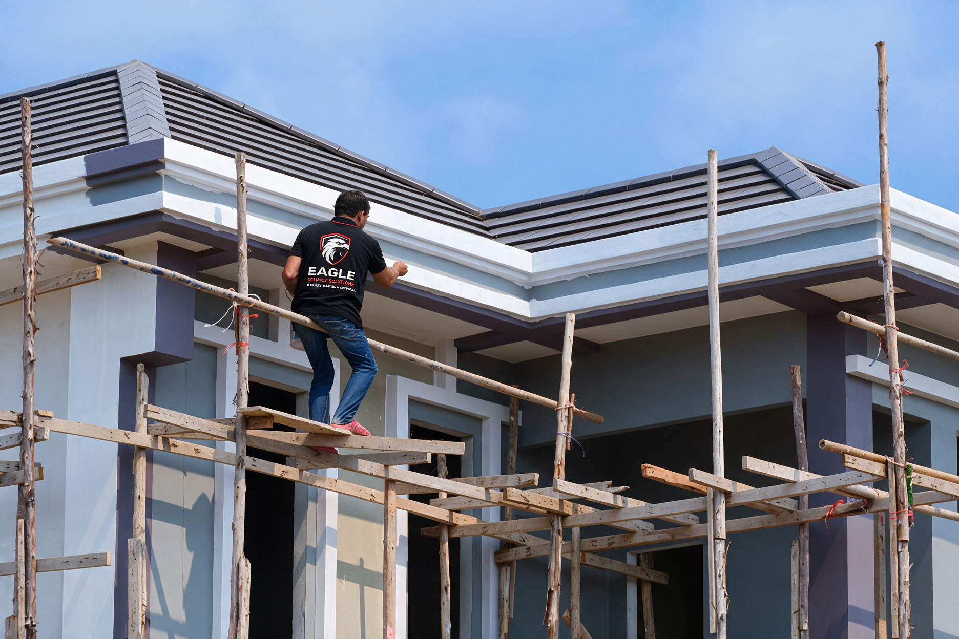 Worker on wooden scaffolding painting the exterior of a modern house with a gray roof.