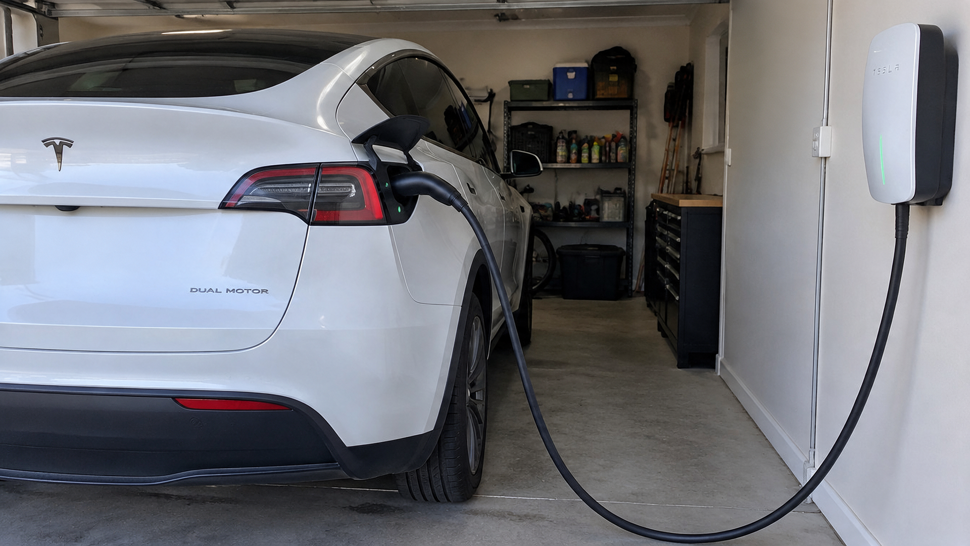 White Tesla parked in a narrow garage, connected to a wall-mounted EV charger.