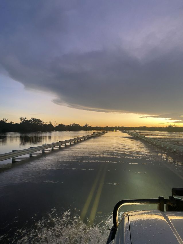 Boat travels across flooded road, leading to distant sunset under a cloudy sky.