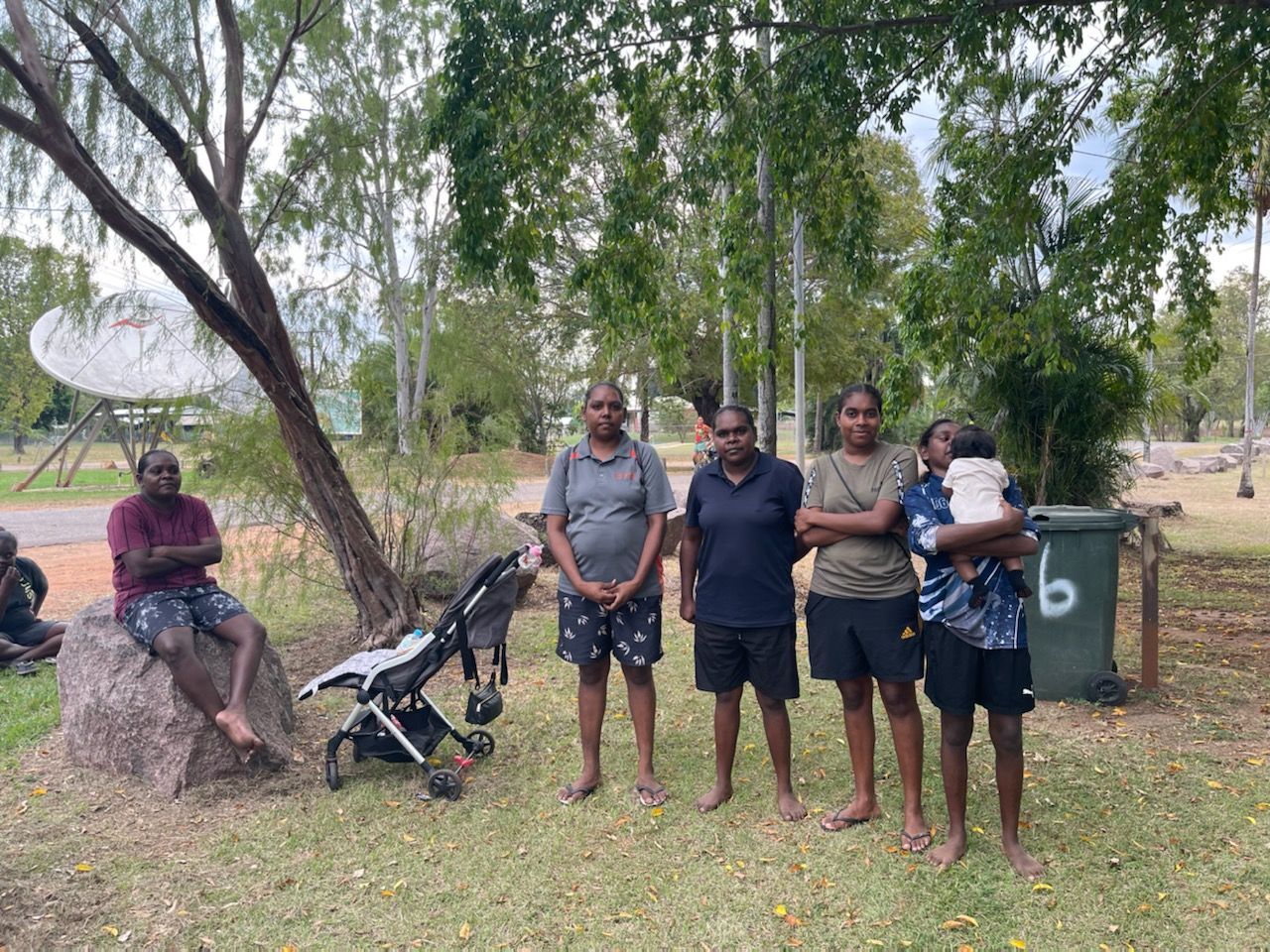 Group of people standing outside near a rock, trees, and a satellite dish. One person is holding a baby.