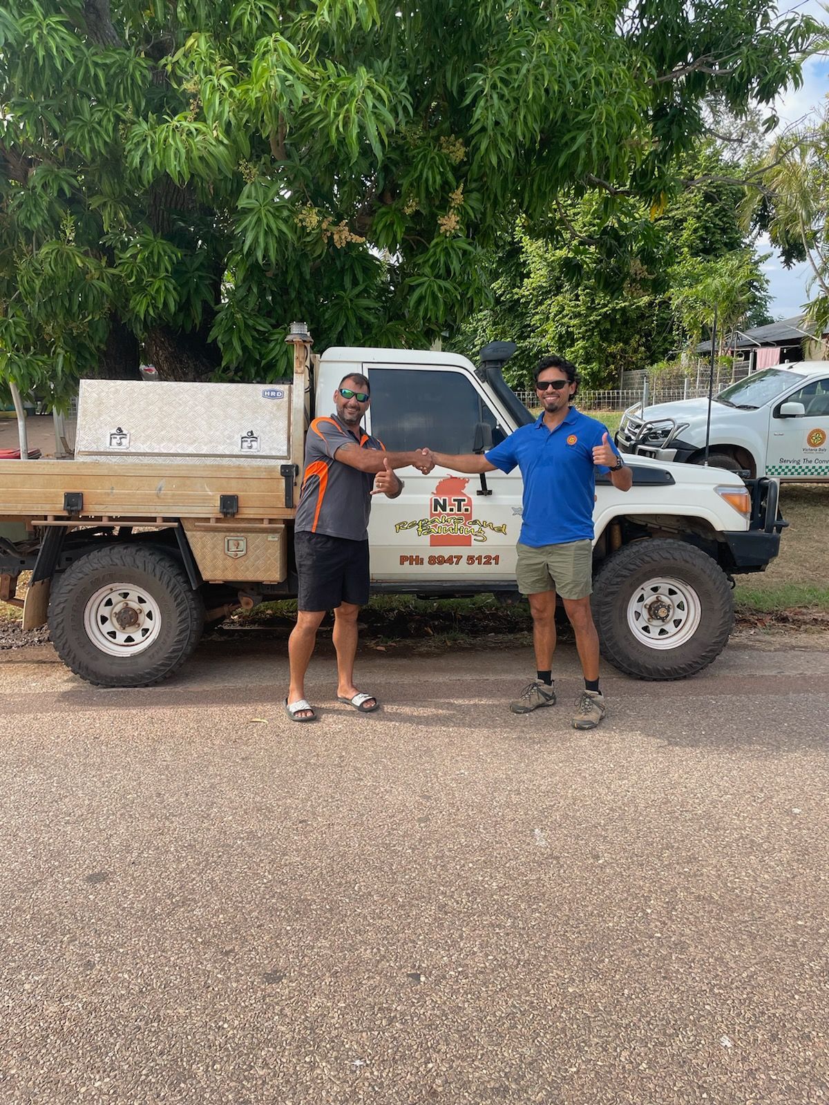 Two men fist bump beside a white utility truck, trees and a parked car in the background.