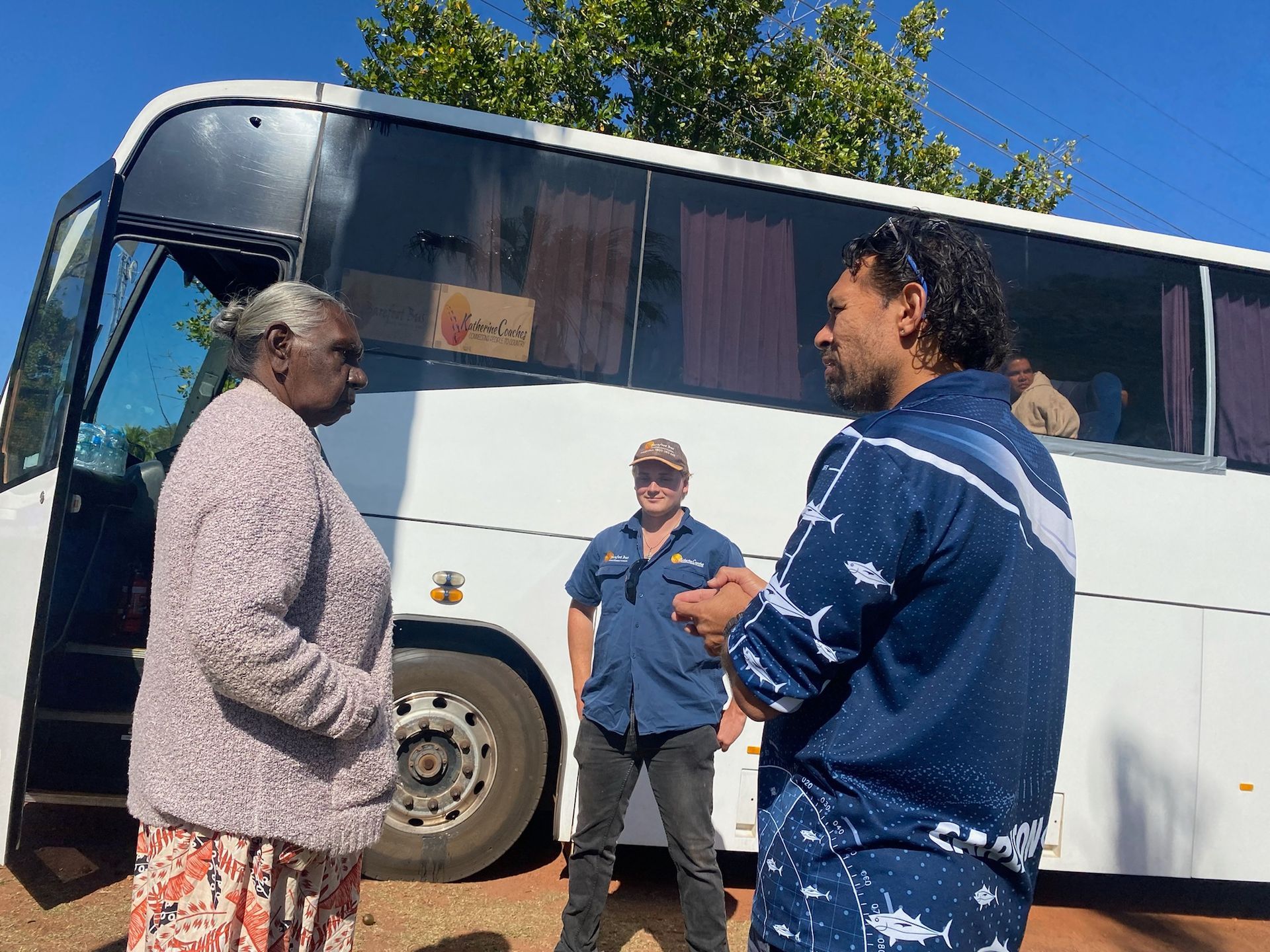 Woman, man, and man in work uniform talking near a white bus outdoors.