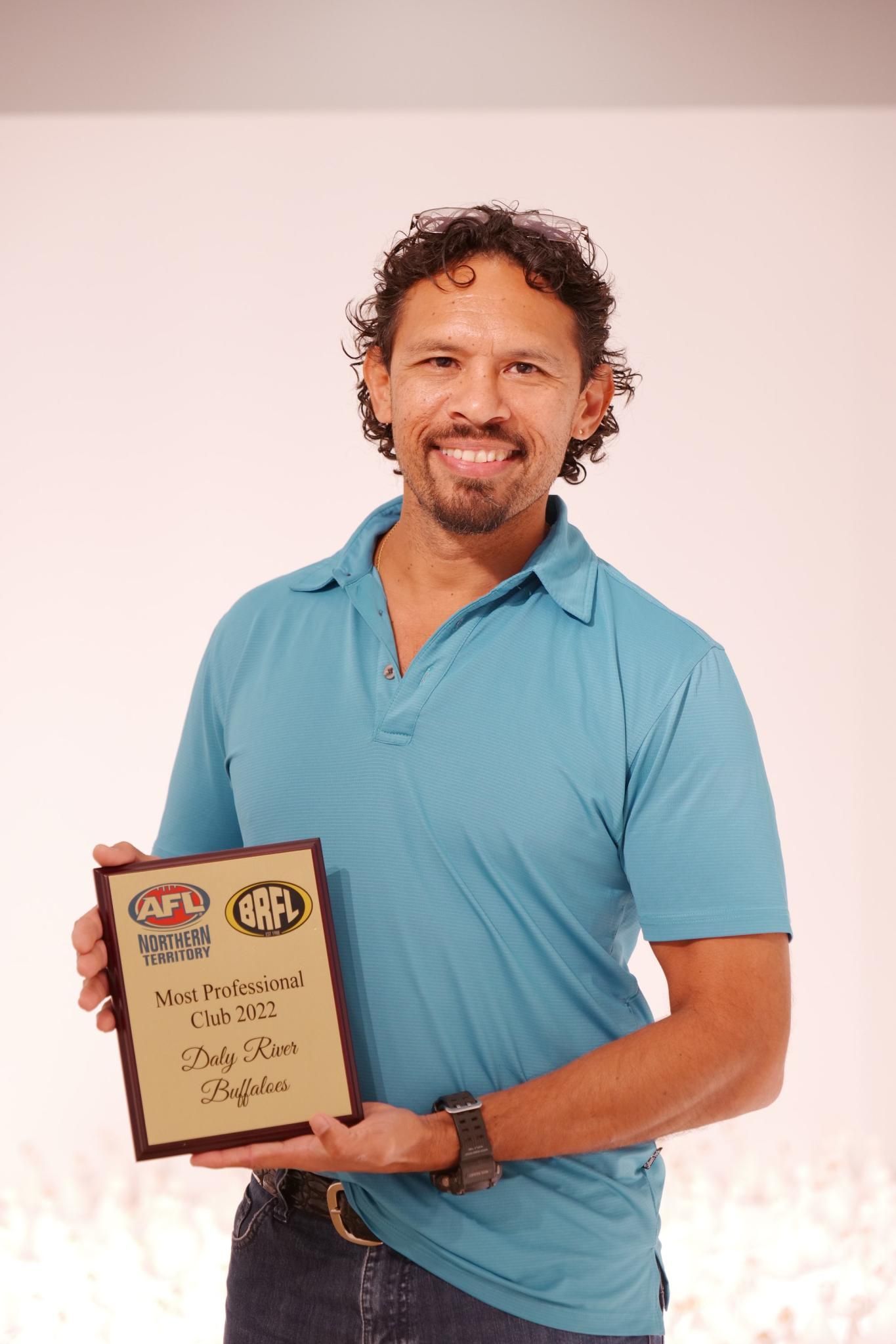 Man in blue shirt smiles, holding plaque with logos. Neutral background.